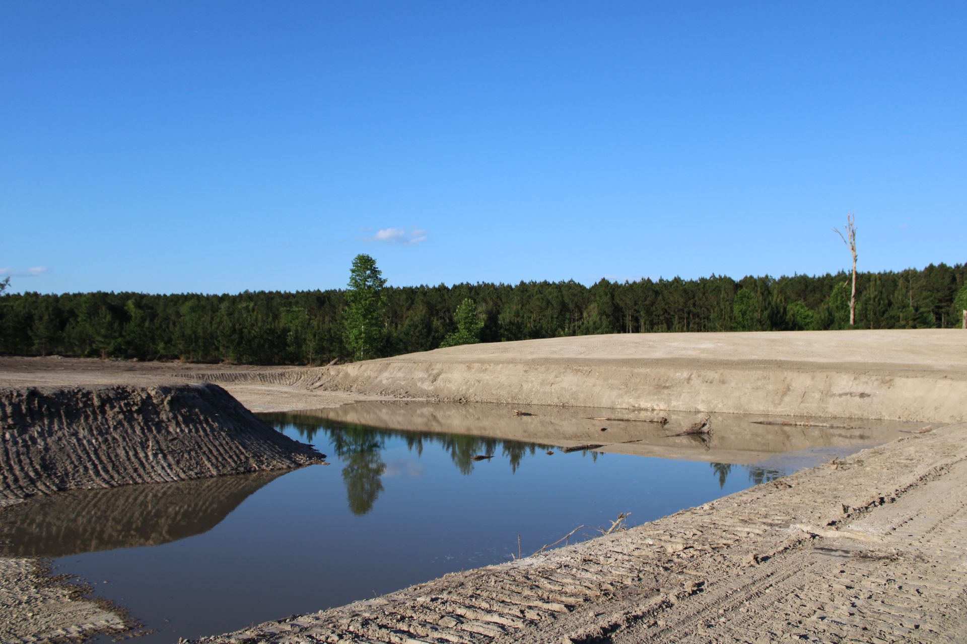 Wide dirt pit with standing water reflecting trees under a clear blue sky