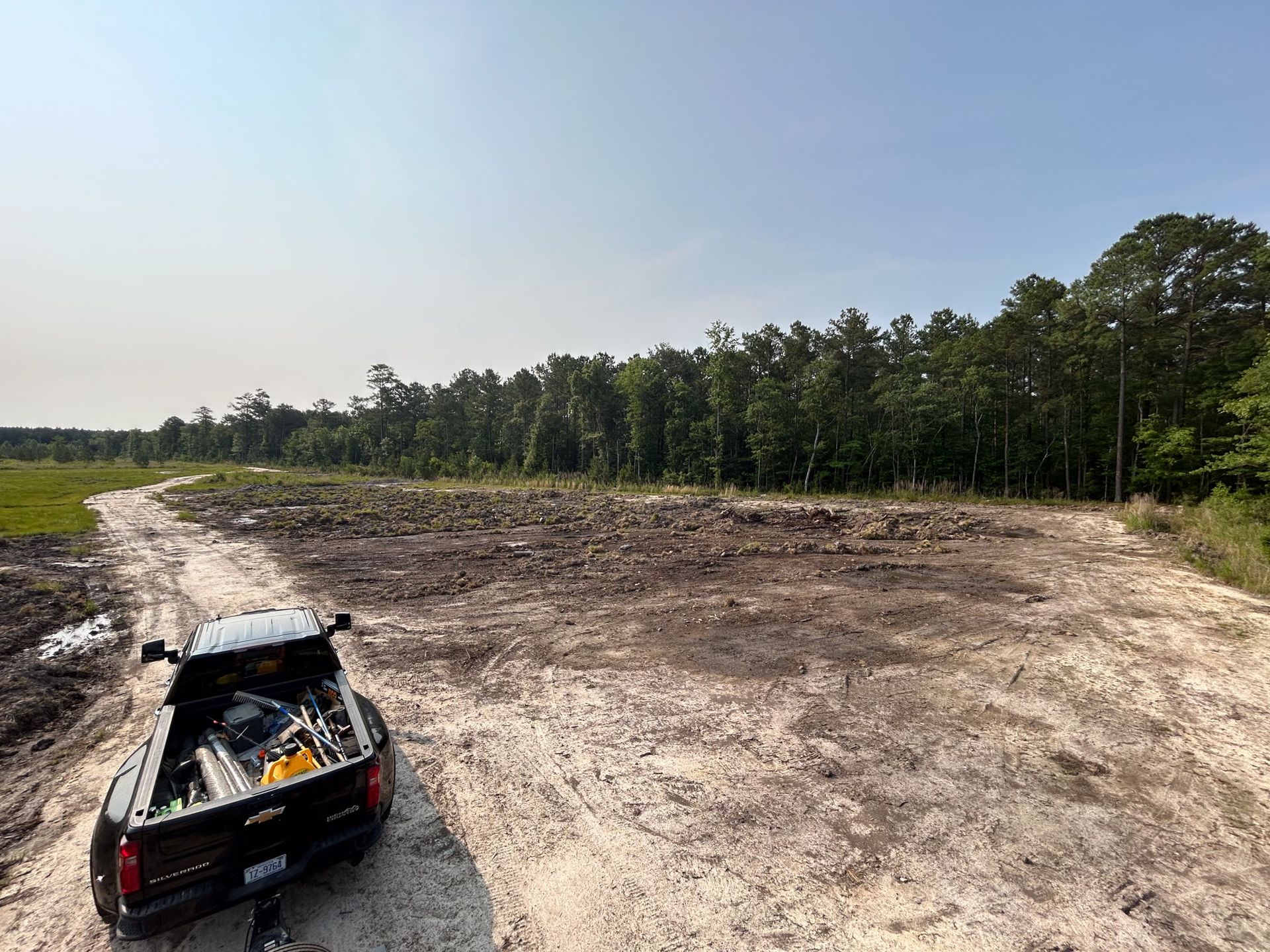 Dirt clearing with truck parked in front, trees in the background, blue sky.