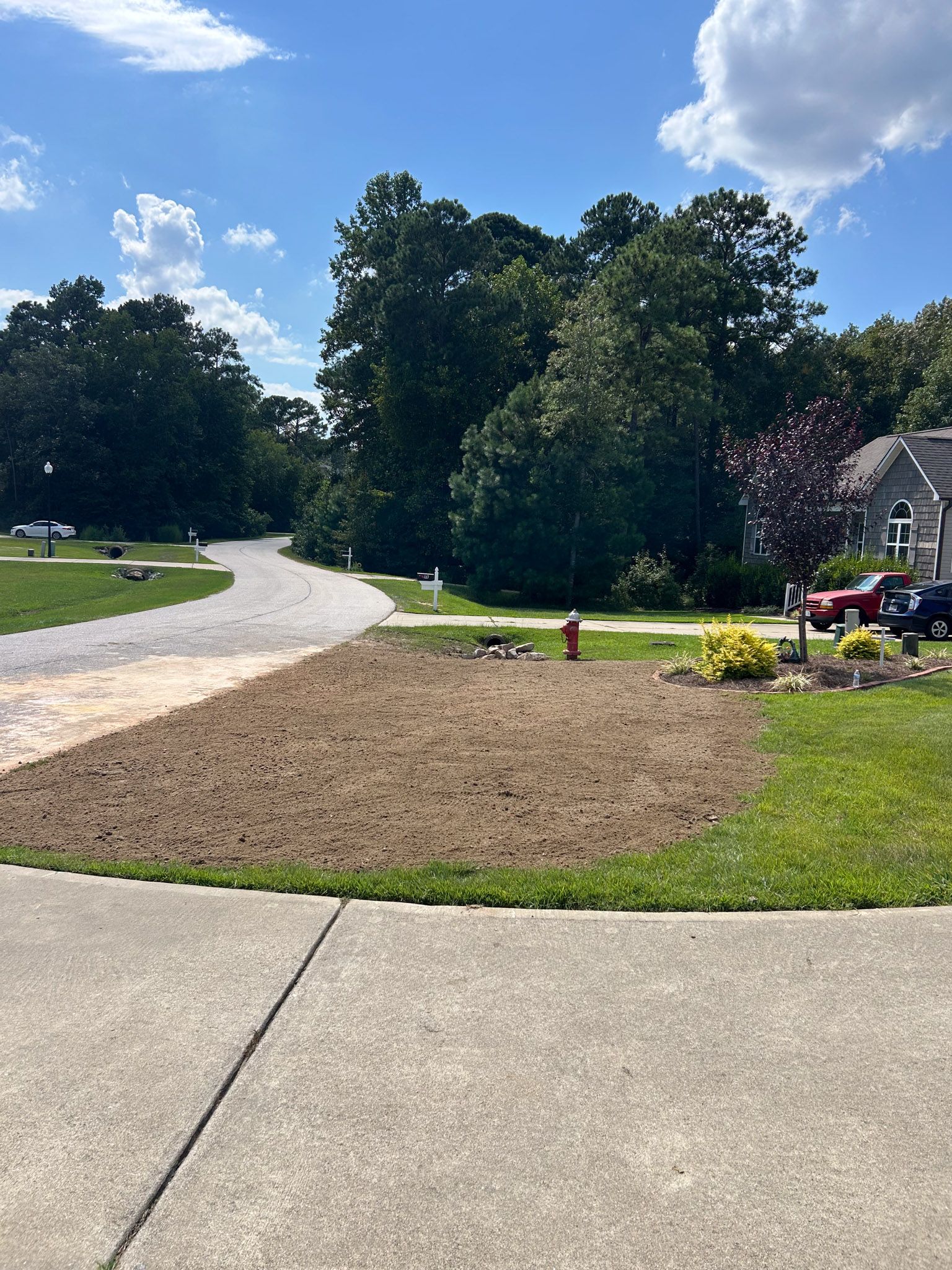 A front yard with newly seeded dirt patch, sidewalk, gravel driveway, and trees under a blue sky.