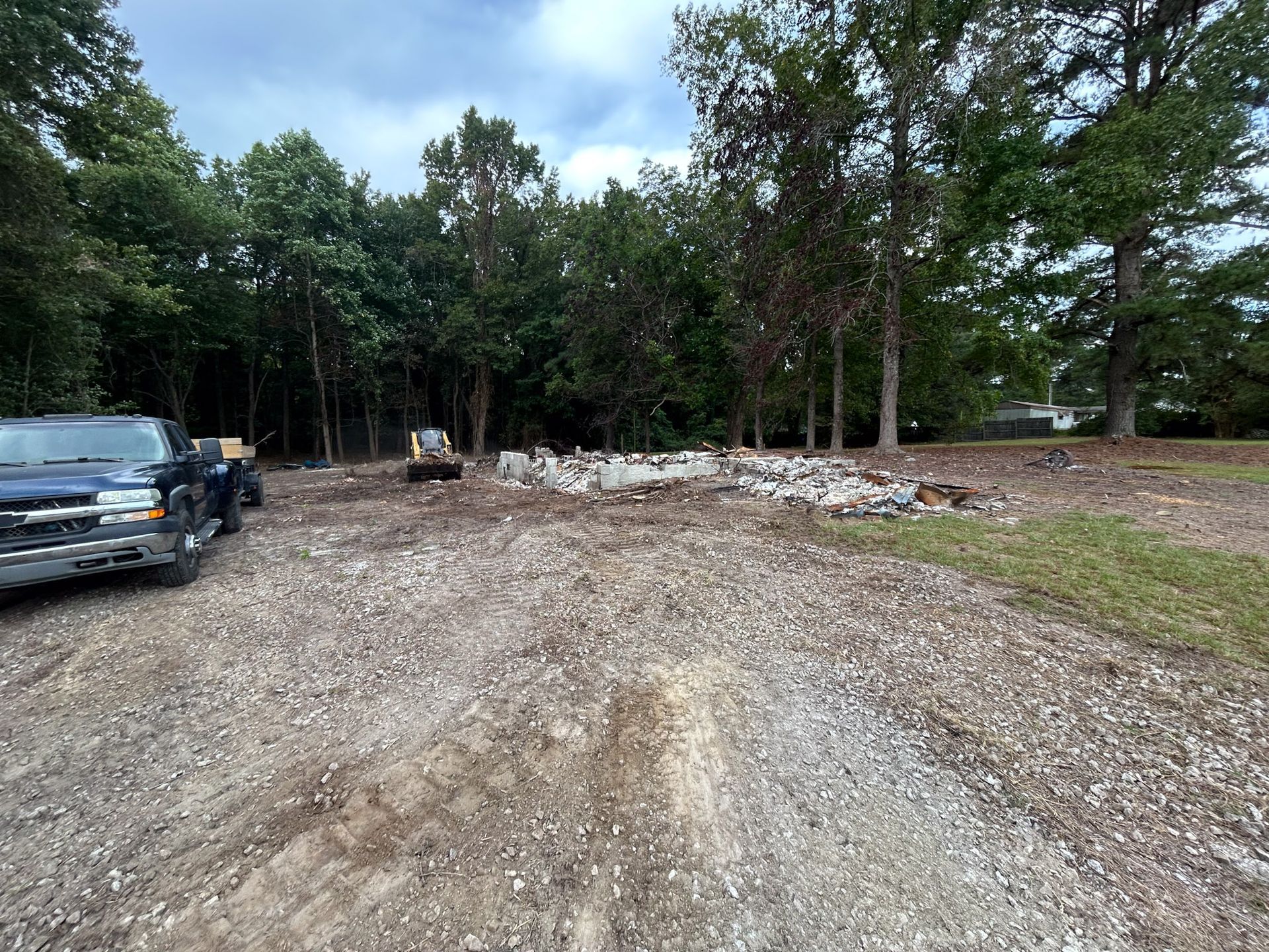 Gravel driveway leads to construction site with debris and a backhoe; trees in the background. A pickup truck is on the left.