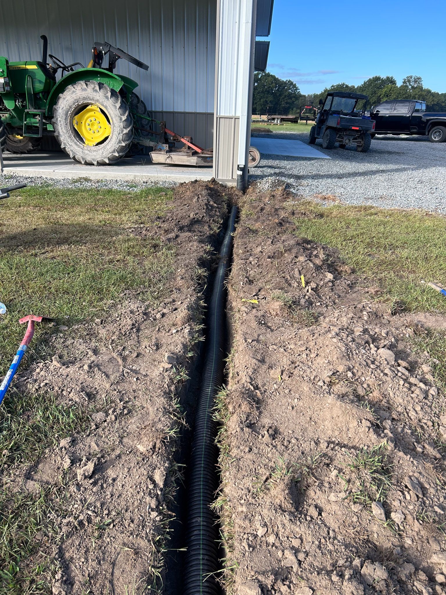 Trench dug next to a building, holding a black drainage pipe. Green tractor and truck in background.