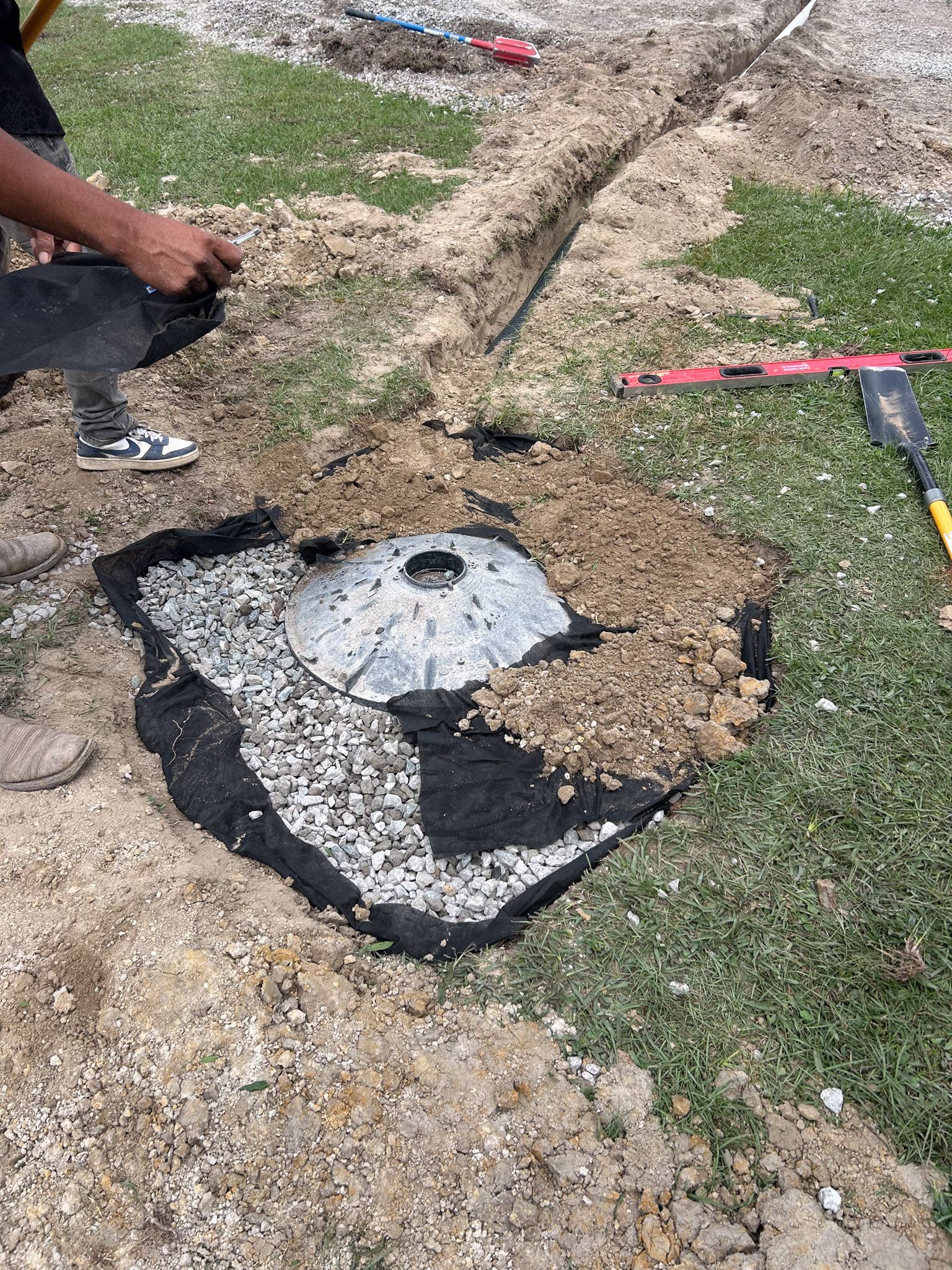 Person installing a drain cover surrounded by gravel, fabric, and soil in a yard.