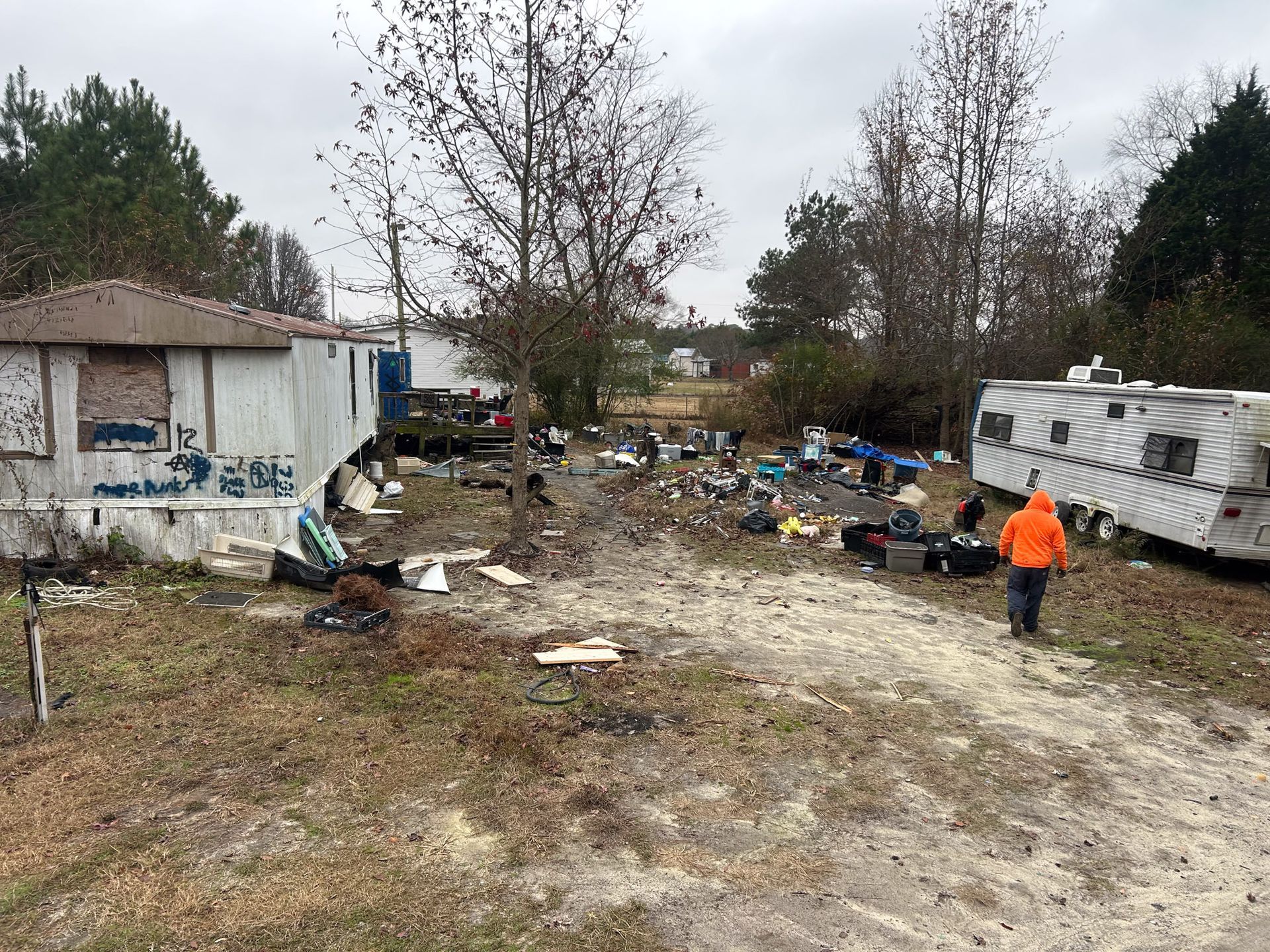 Yard with debris, damaged structures, and a person in orange jacket. Overcast day.