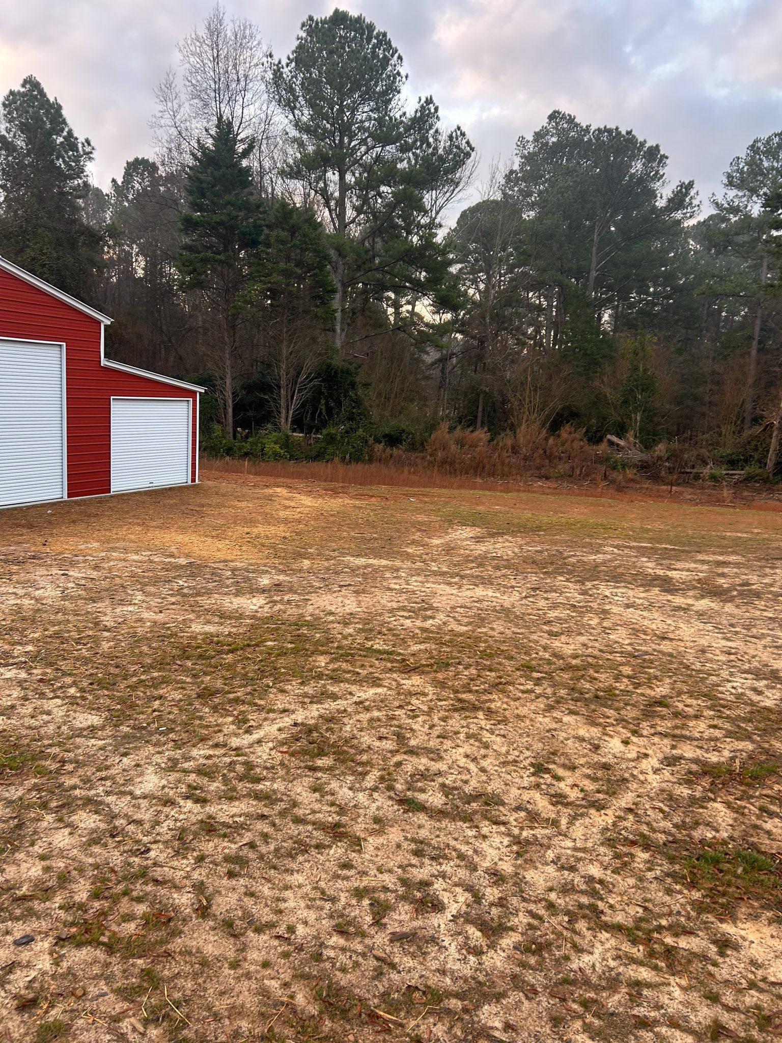 Red building with white doors next to a brown field and a forest of green trees.