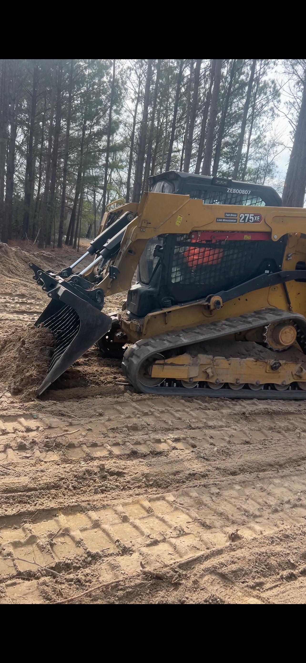 A yellow skid steer with a grapple attachment is on muddy ground, trees in the background.