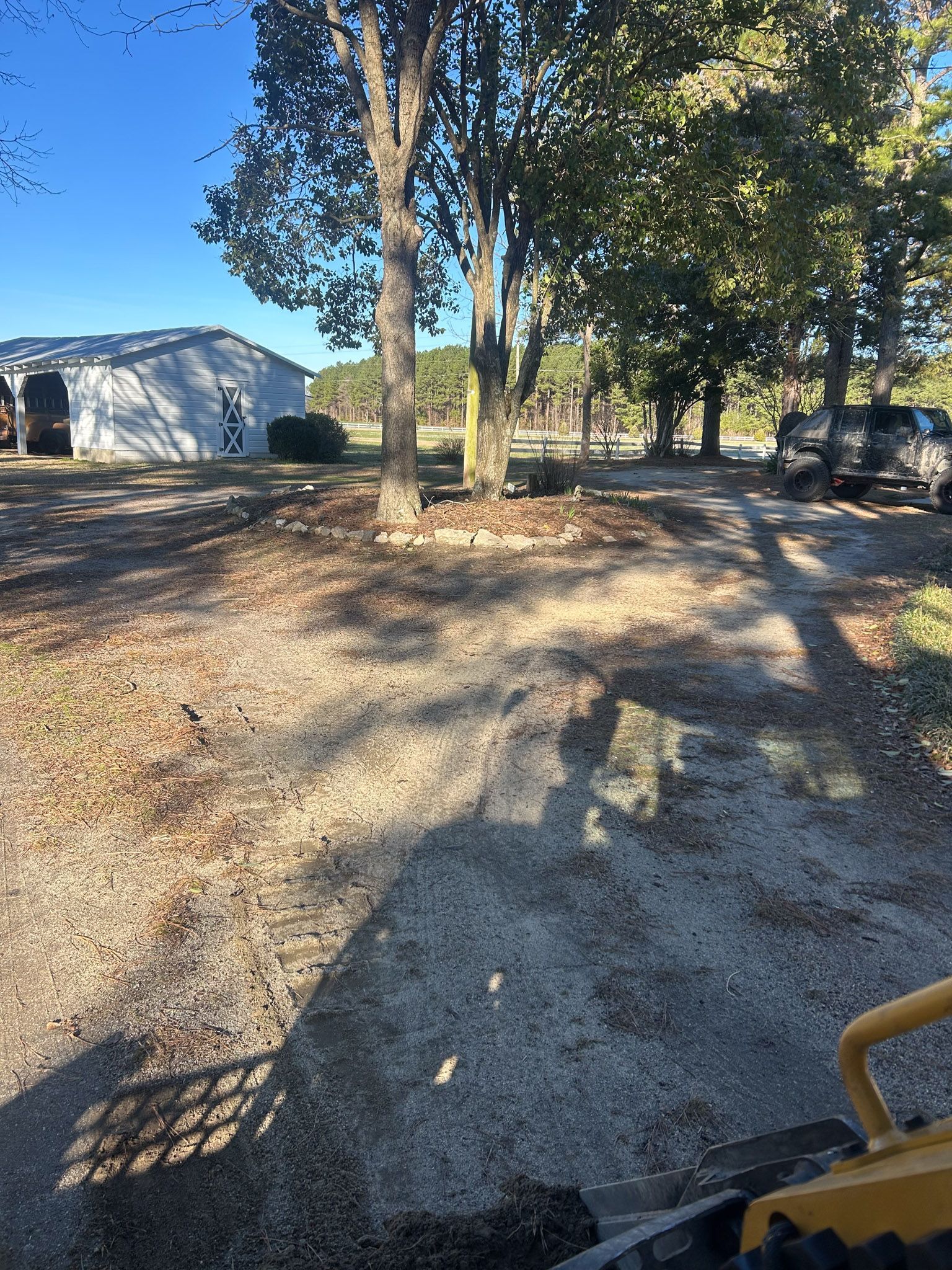 Gravel driveway leading to a barn, with a tractor in the foreground. Trees and a vehicle are in the background.