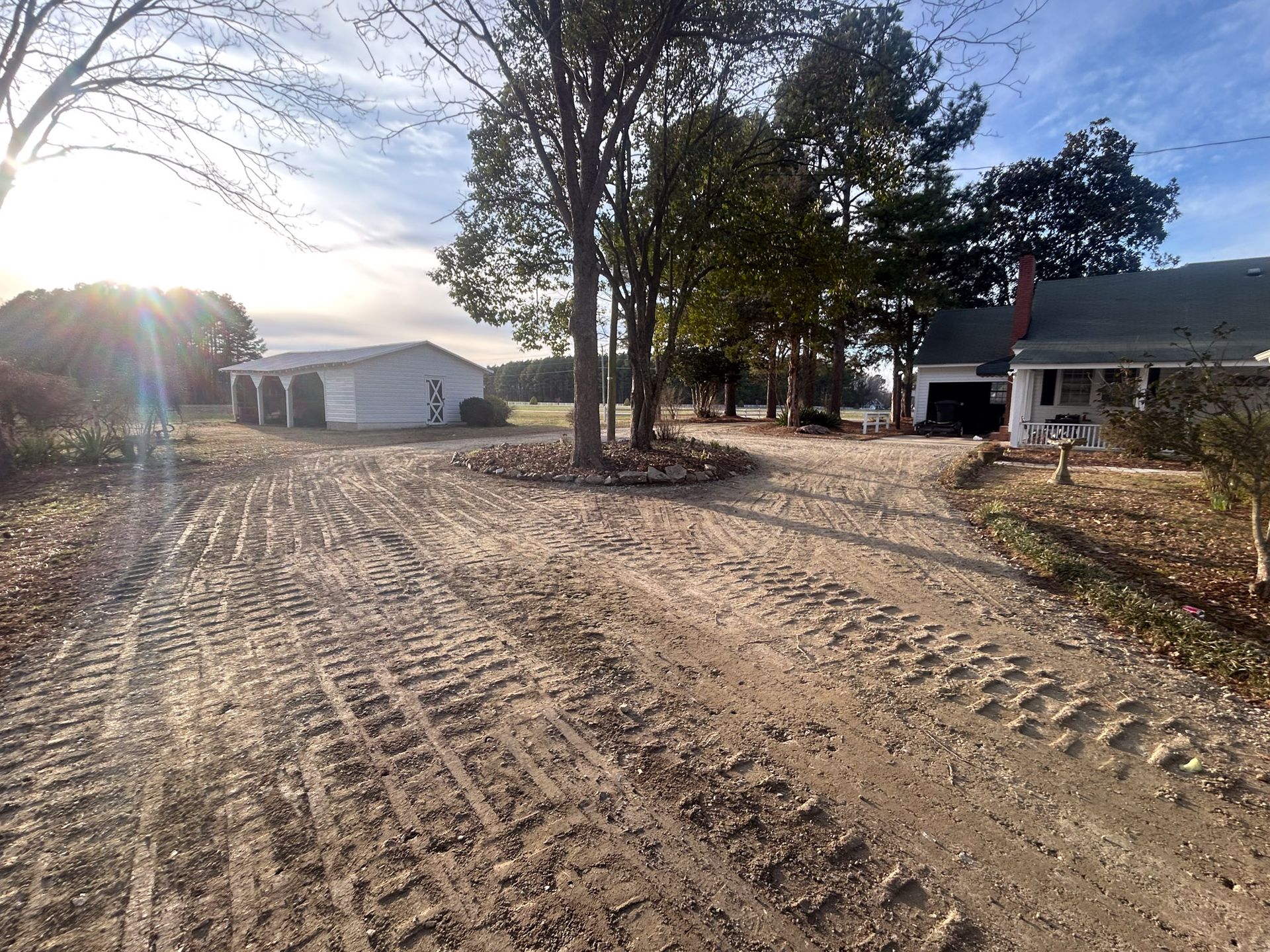 Dirt driveway with tire tracks, trees, a white garage, and a house under a cloudy sky.