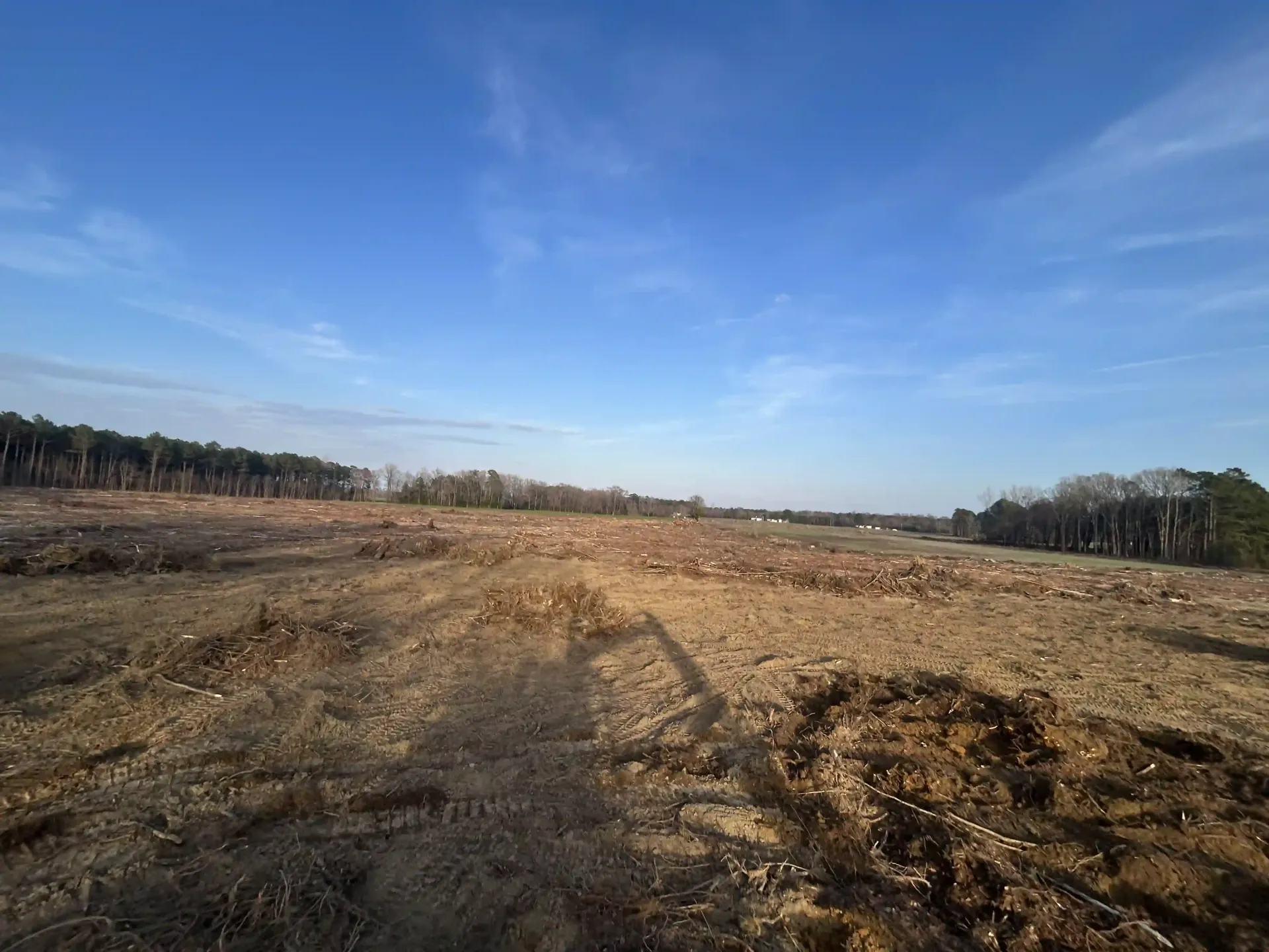 Wide barren dirt field under a blue sky, with trees lining the horizon.