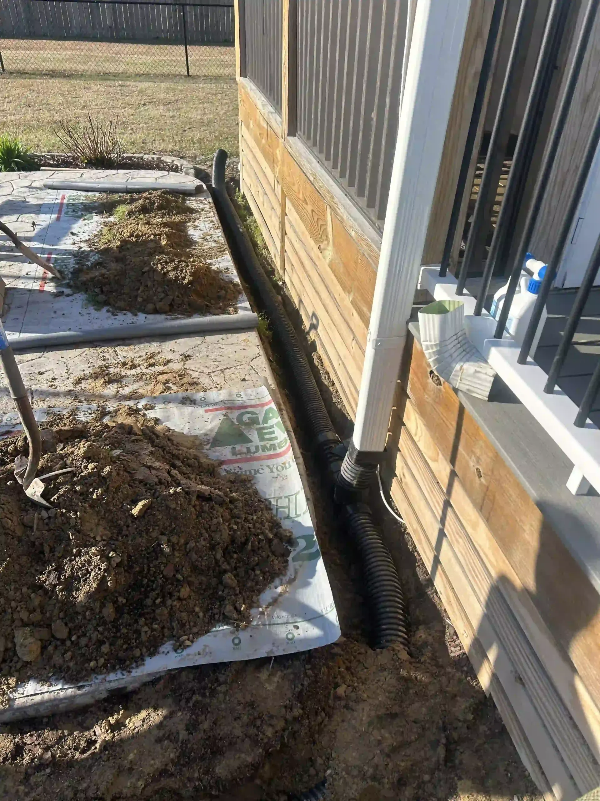 Mulch pile and torn landscaping fabric beside a house foundation and fence, with a drainage pipe along the wall