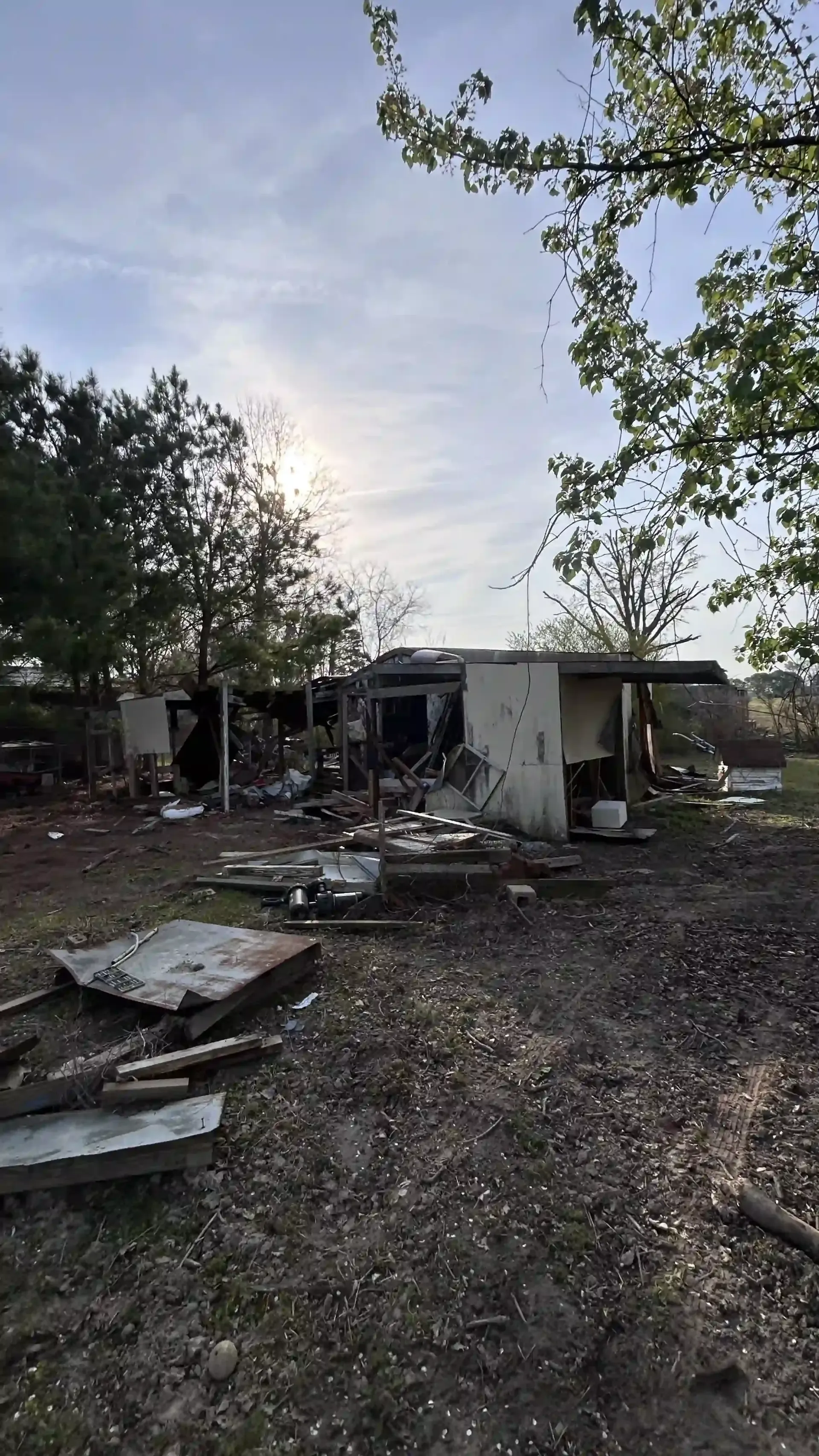 Damaged house with debris scattered in yard at dusk, surrounded by trees and an overcast sky