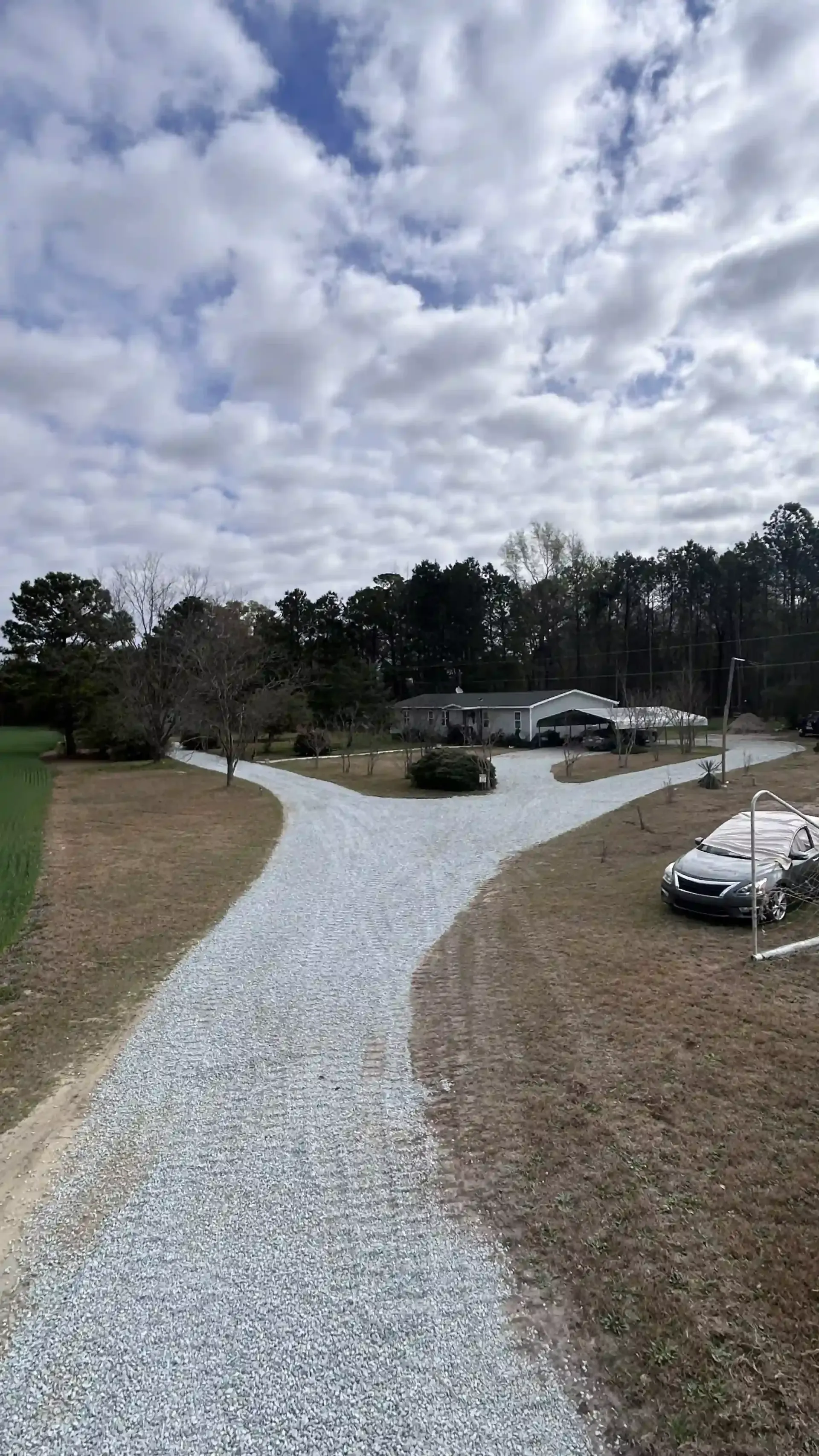 Gravel driveway leading to a house, with parked cars and trees under a cloudy sky.