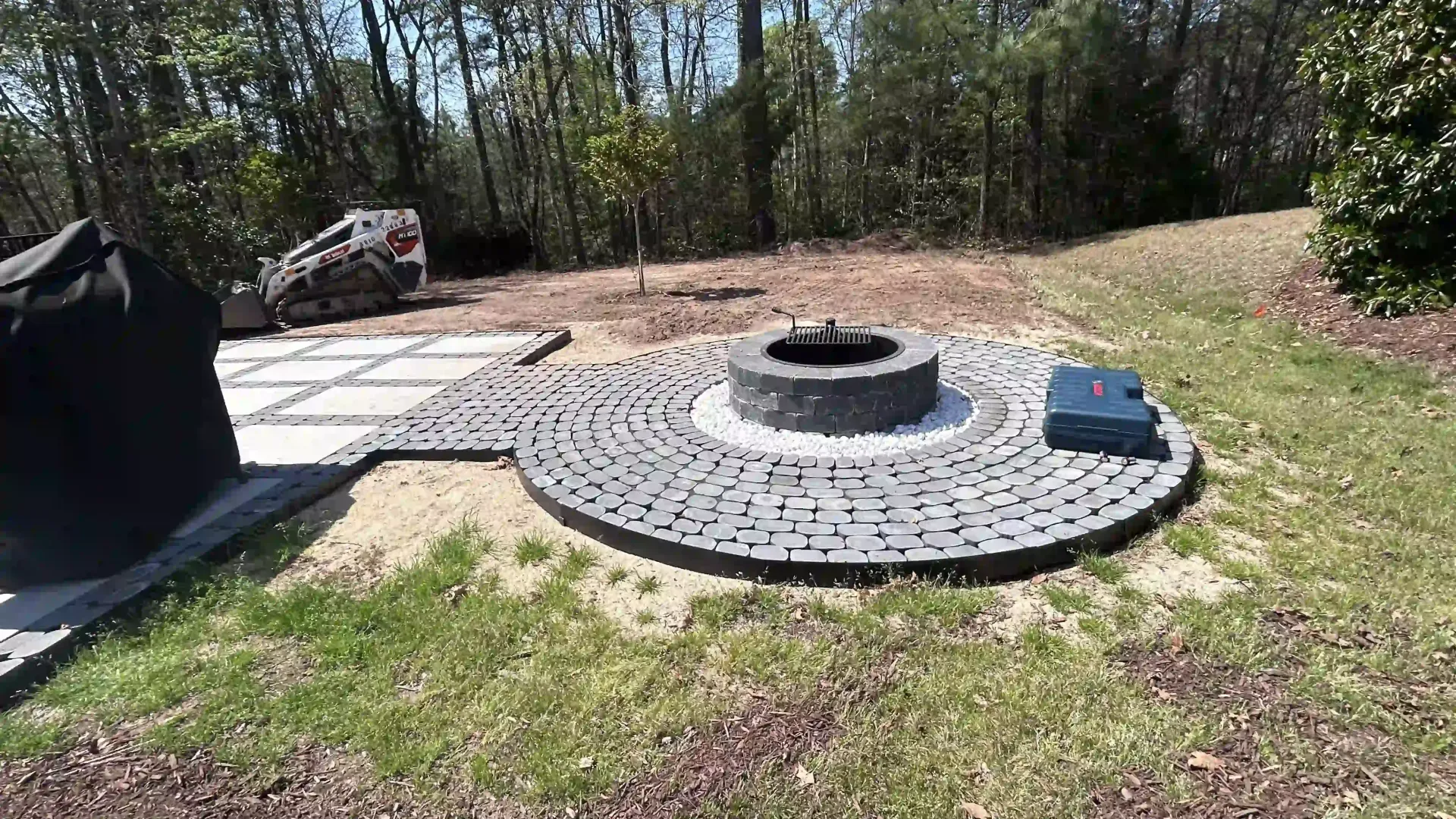 Outdoor patio with circular stone fire pit and paver seating area beside grass and trees
