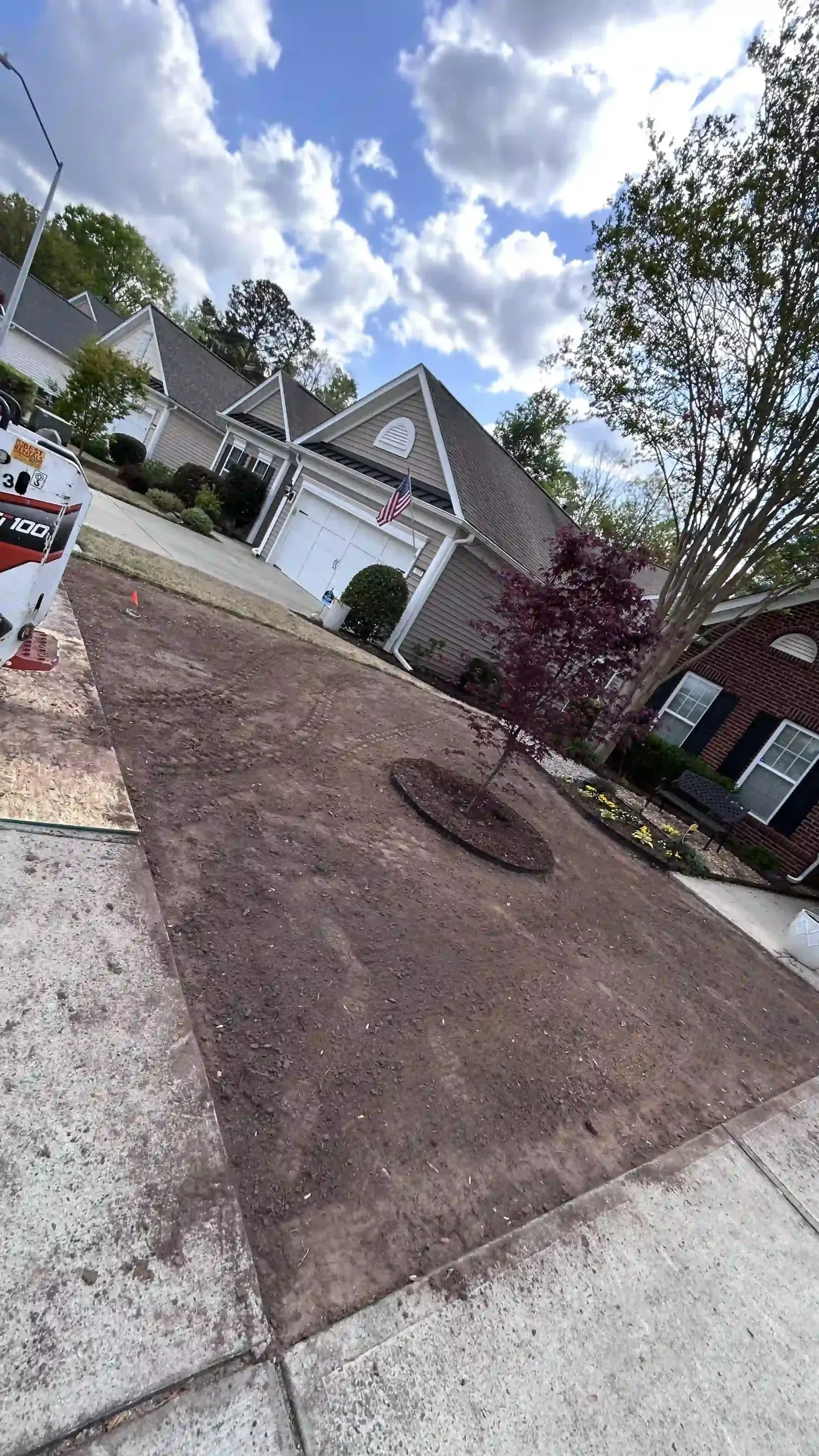 Residential driveway with a large dirt patch beside a white house under a cloudy sky.