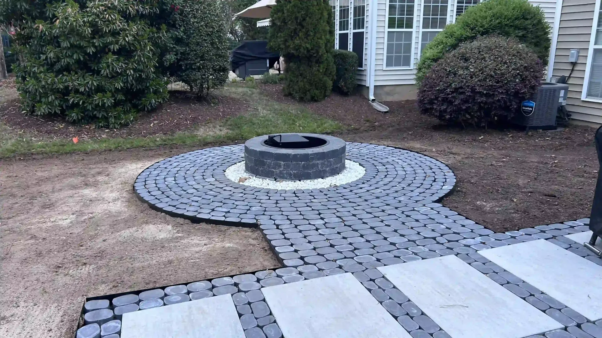 Backyard patio with a round fire pit and gray paver walkway leading to a house entrance