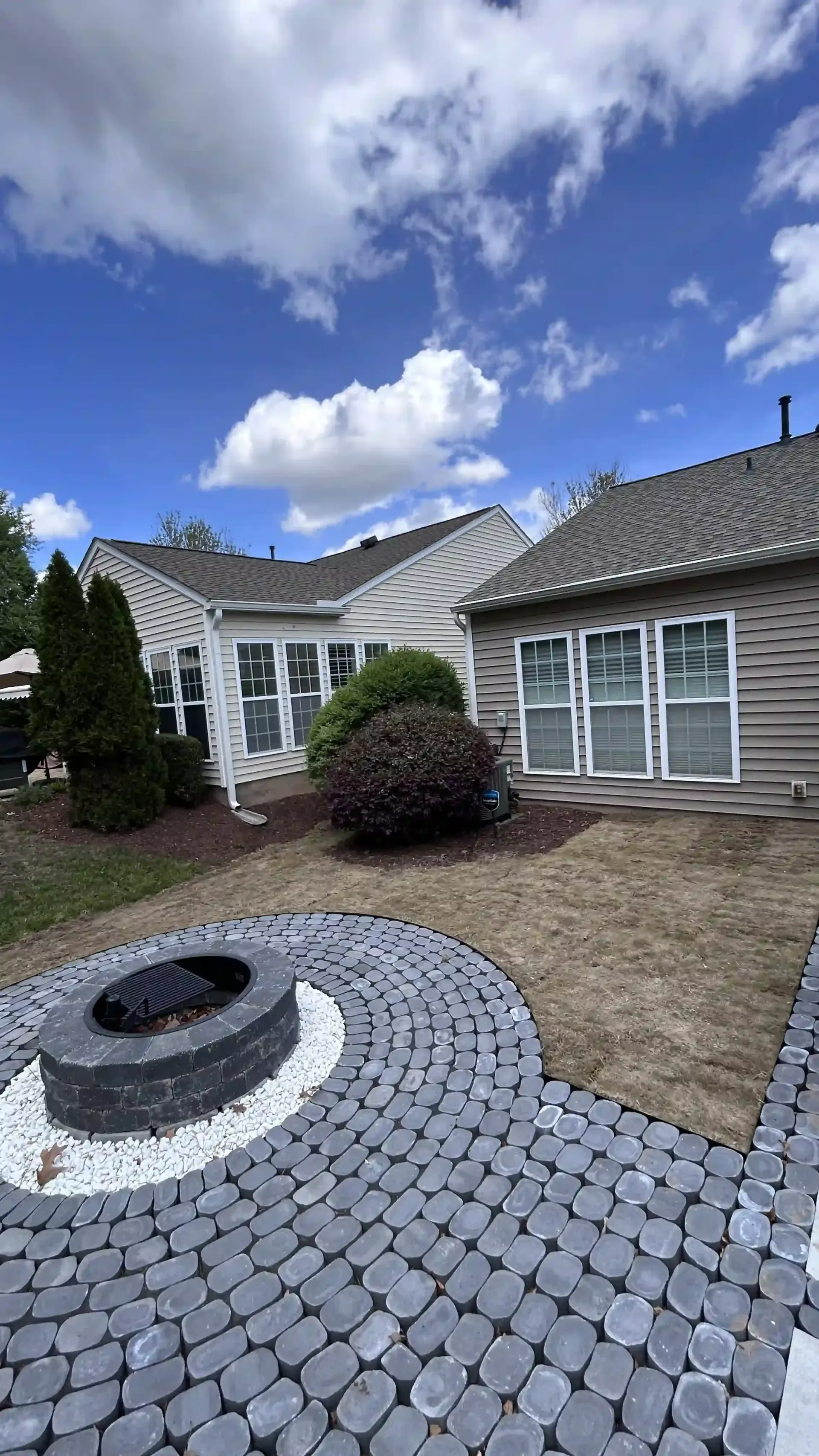 Backyard patio with circular stone fire pit, white house, and cloudy sky