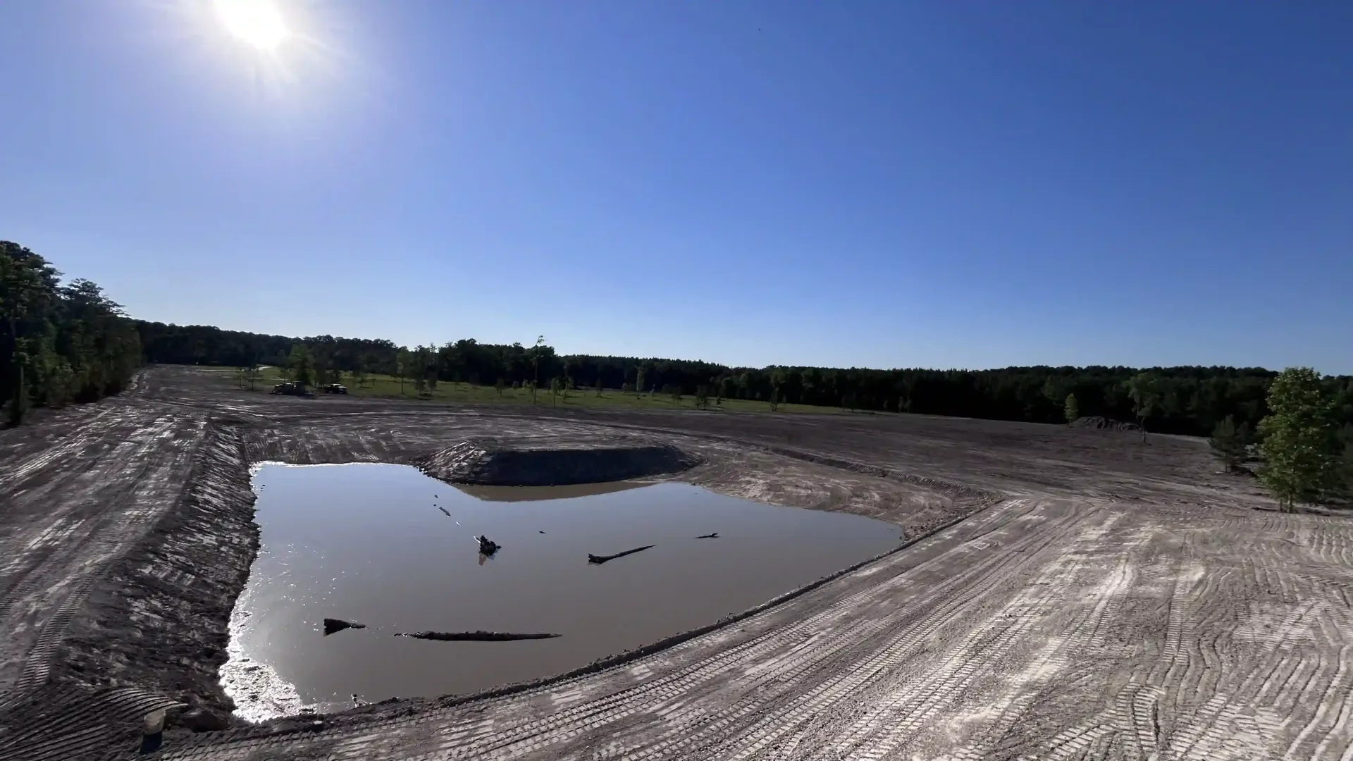 Wide muddy excavation pit with standing water under a clear blue sky, bordered by dirt and trees