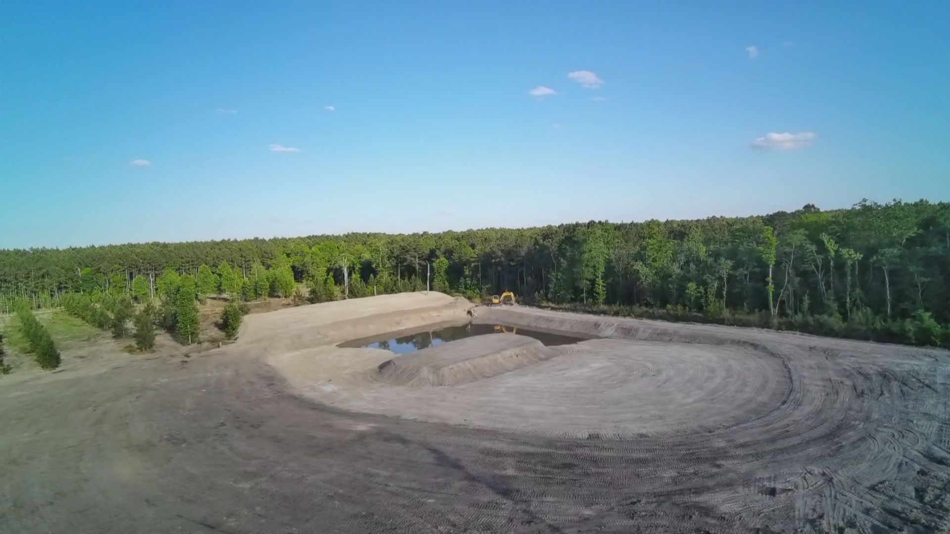 Aerial view of a dirt construction site surrounded by green forest under a blue sky.