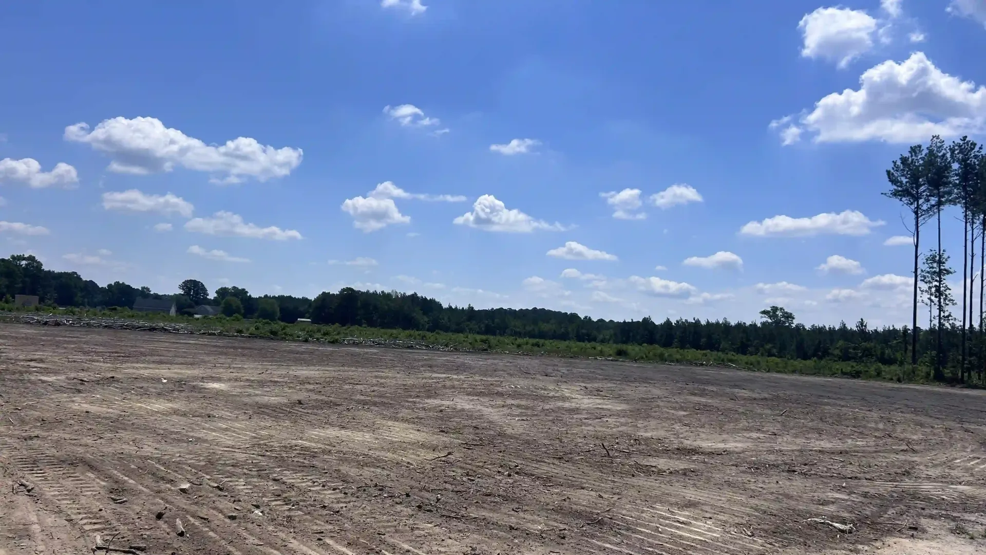Wide cleared dirt field under a bright blue sky with scattered clouds and a tree line in the distance