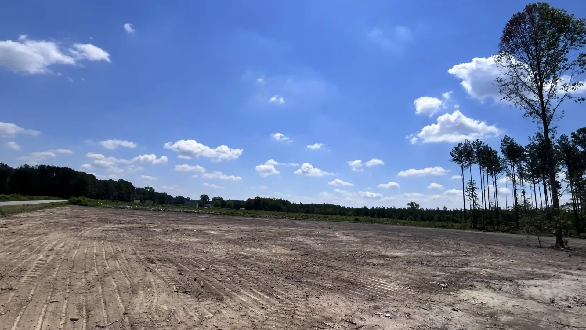 Cleared dirt field under a bright blue sky with clouds and trees on the horizon