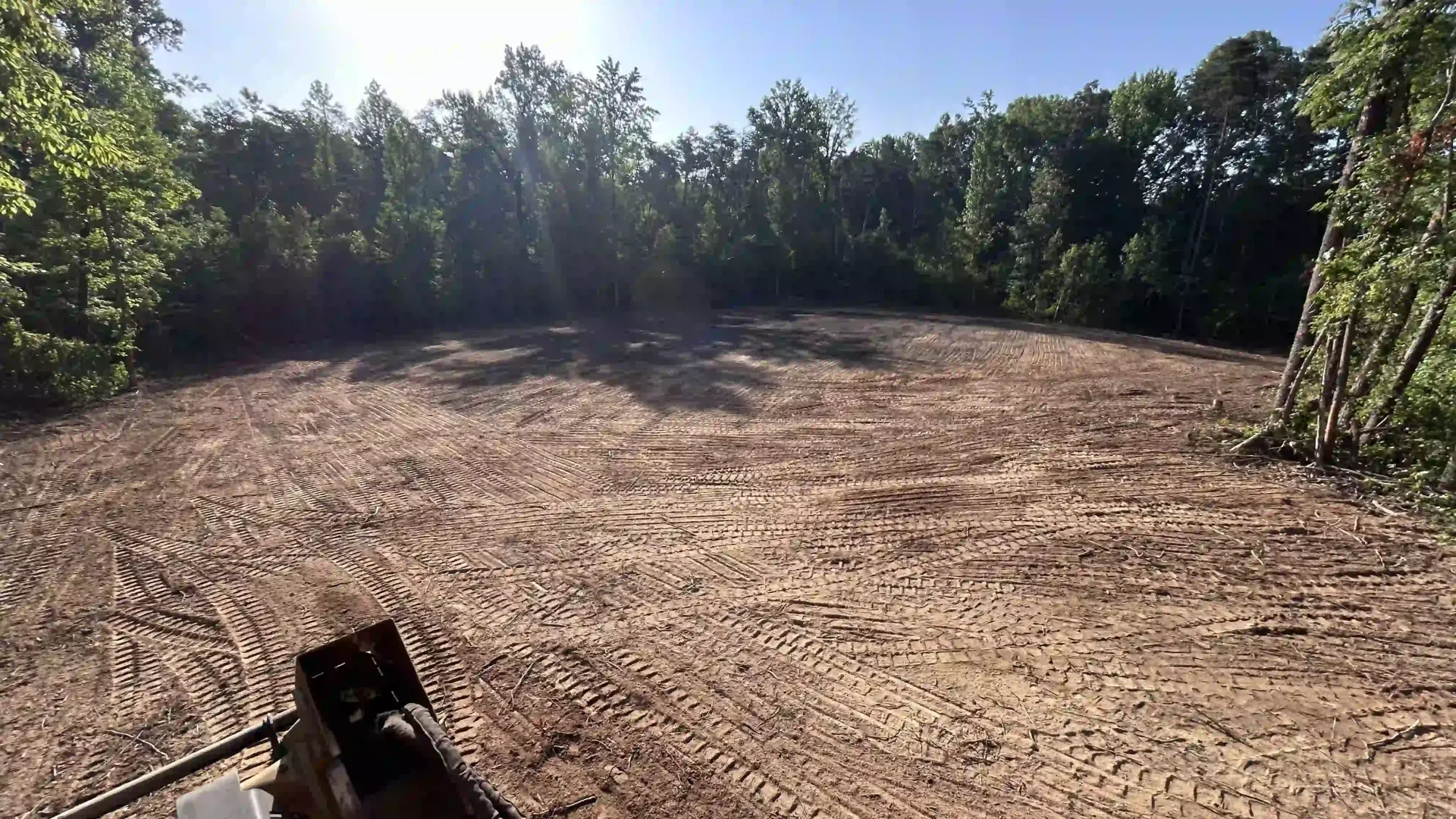 Cleared dirt lot bordered by trees under bright sunlight, with a heavy machine visible at bottom left.