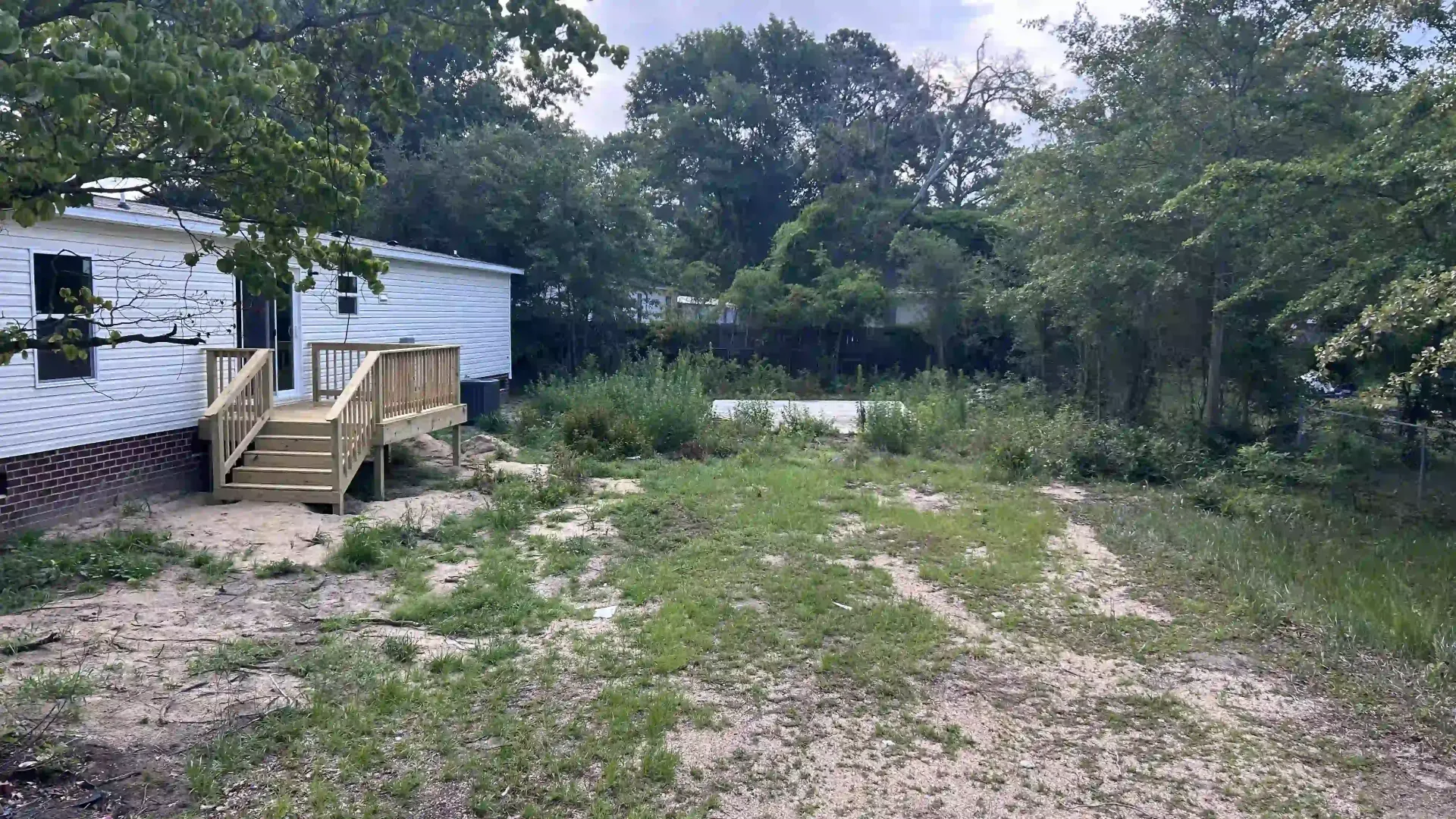 Gravel yard beside a white mobile home with a small wooden porch, surrounded by trees and greenery.