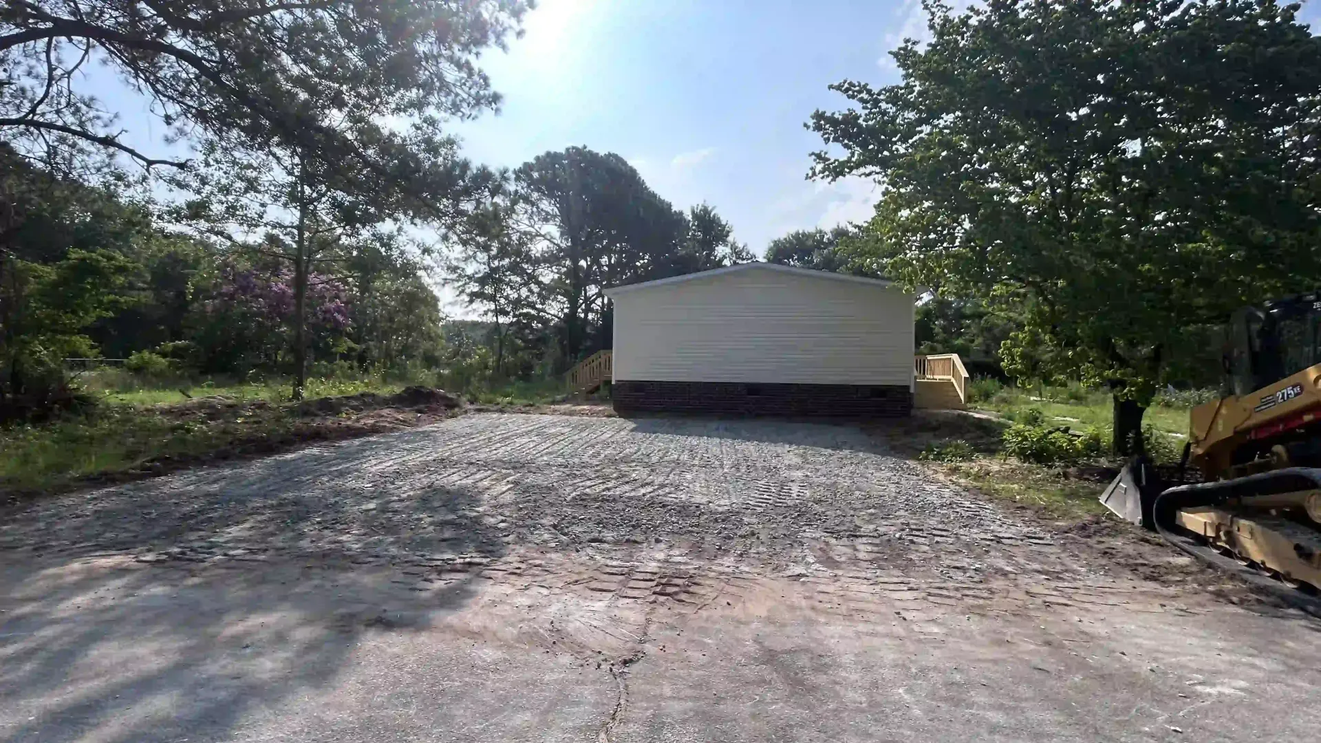 Gravel clearing with a white trailer or container centered, trees and construction equipment on the sides.