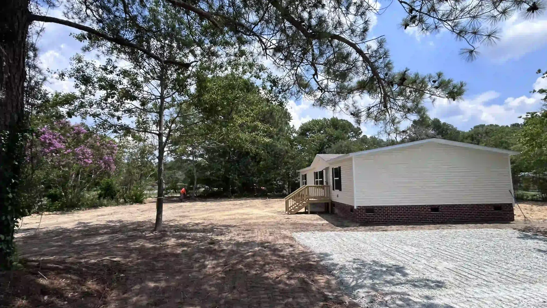 White mobile home on gravel lot under trees, with a small porch and blue sky in the background