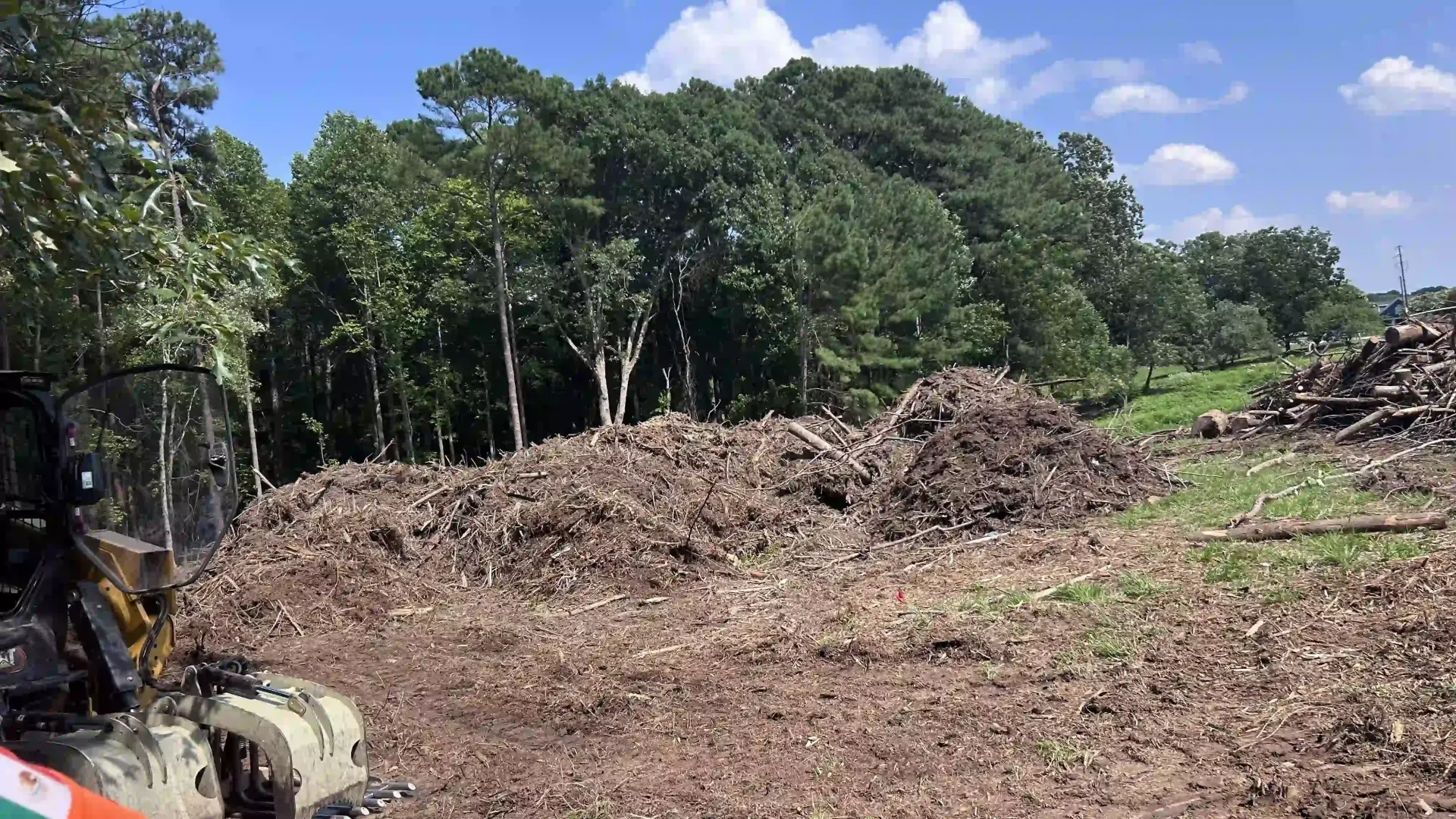 Cleared dirt pile in a wooded area under a blue sky, with an excavator at the left edge