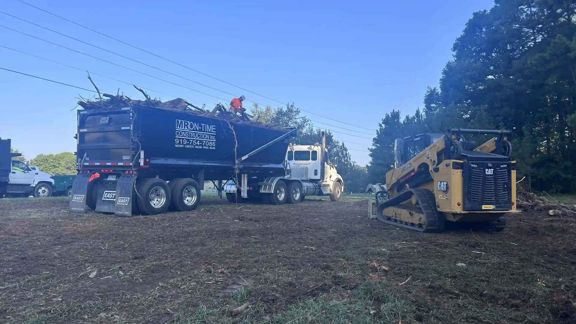 Black garbage truck and yellow wood chipper in a wooded worksite under a blue sky