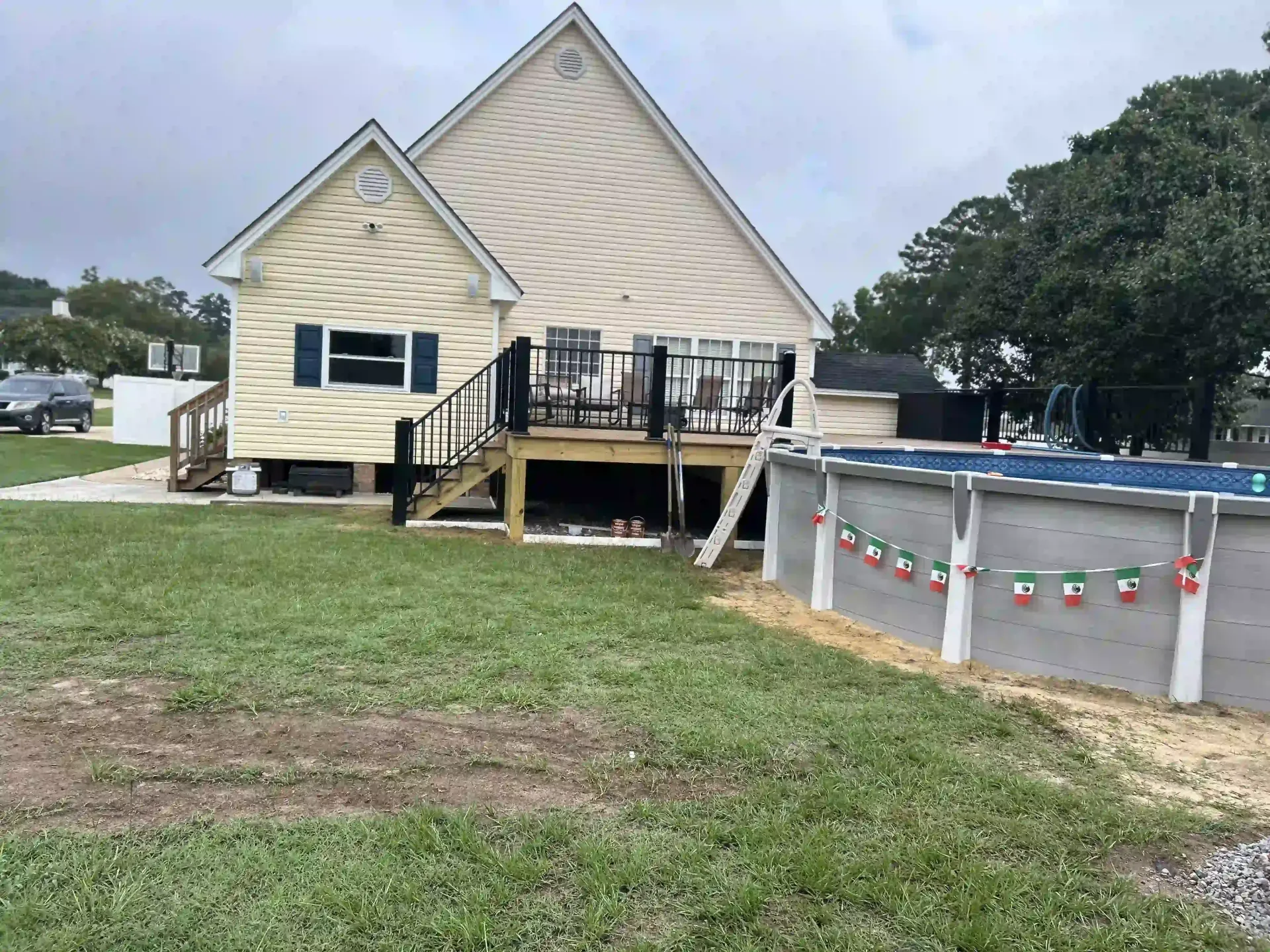 A beige house with a raised deck beside a above-ground pool and grassy yard.