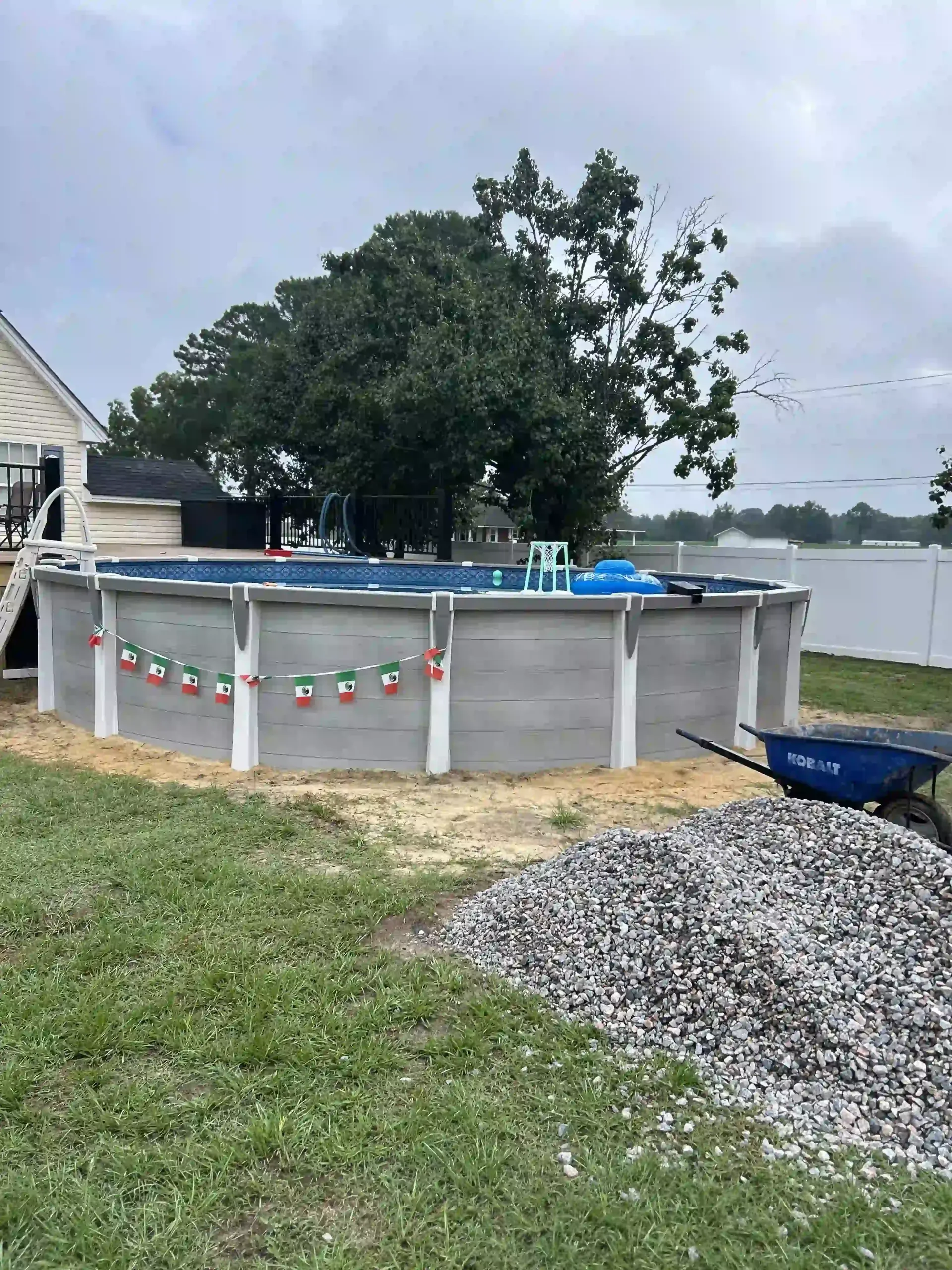 Above-ground pool in a backyard beside a house, with gray siding, gravel, and a tree nearby.