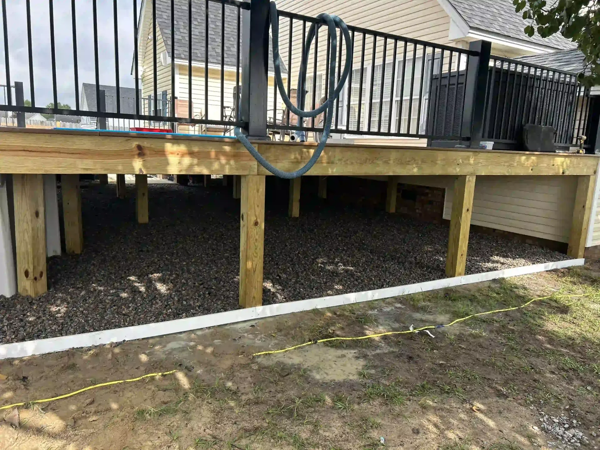 Raised wooden deck with black railing and support posts over gravel, beside a house.