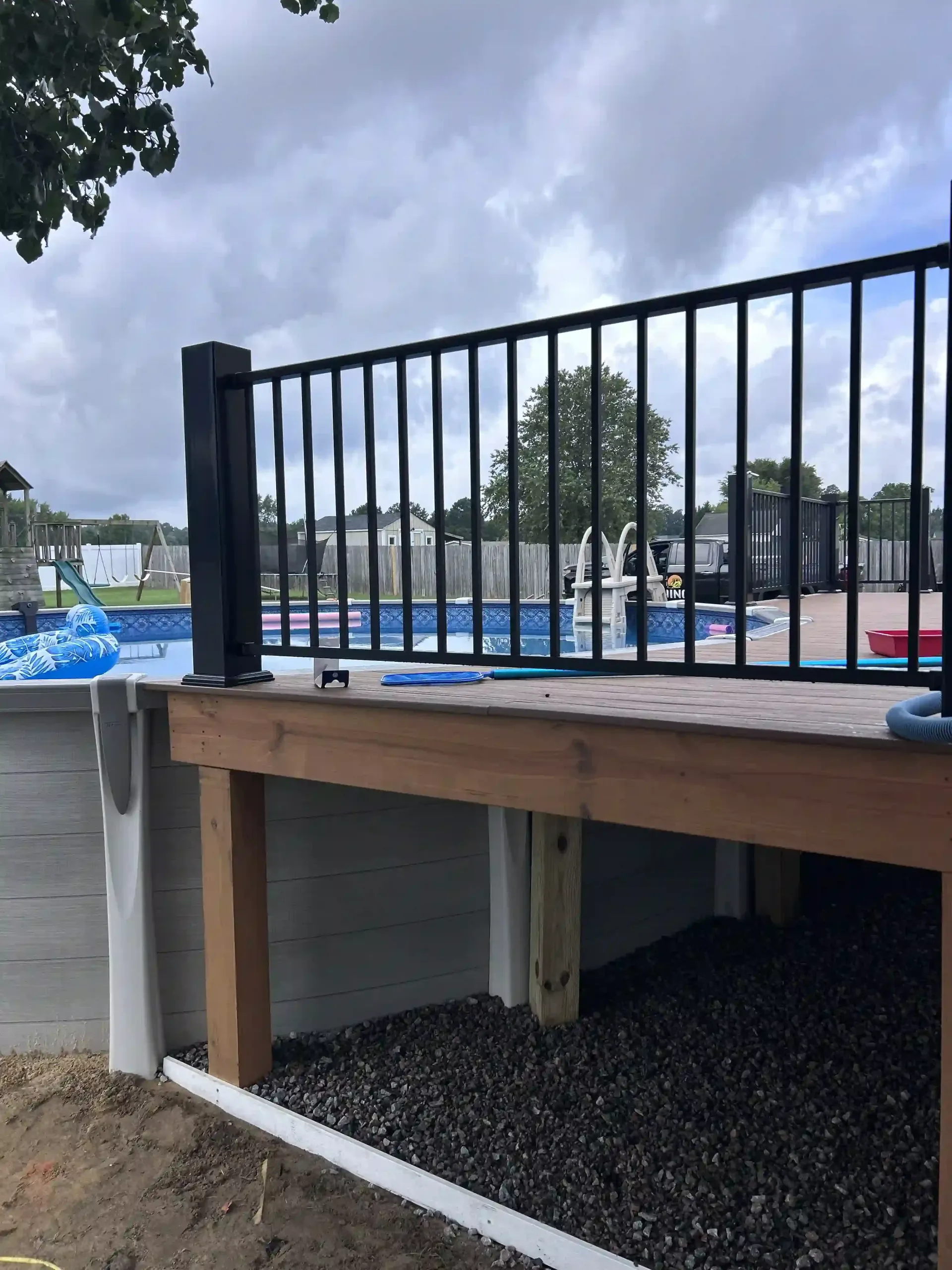 Wooden platform with black railing by a waterfront, with blue tarps and trees in the background