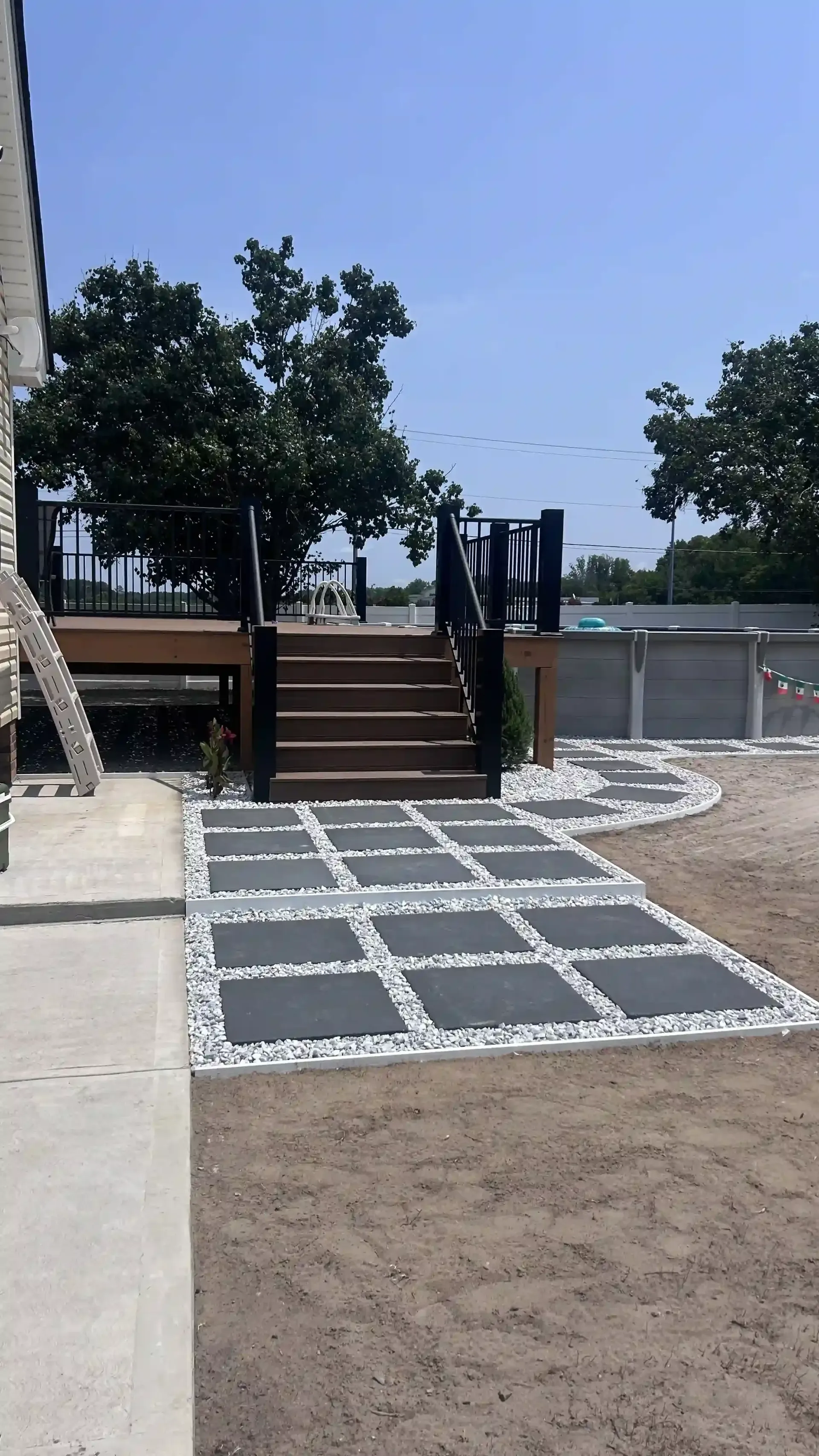 Backyard with stepped wooden deck, black railings, and a square paver path in gravel under a clear sky