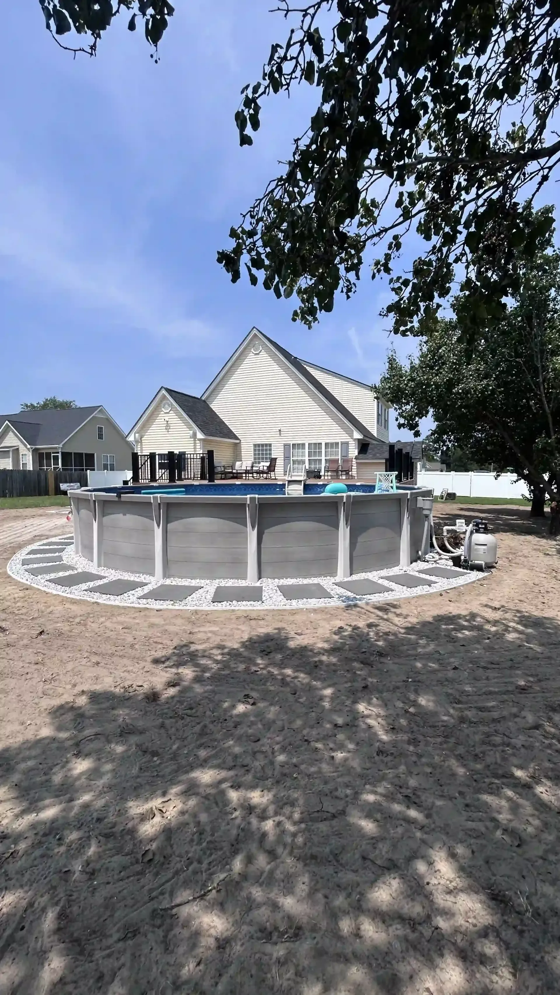 Backyard above-ground pool beside a white house, with people gathered on the deck under trees