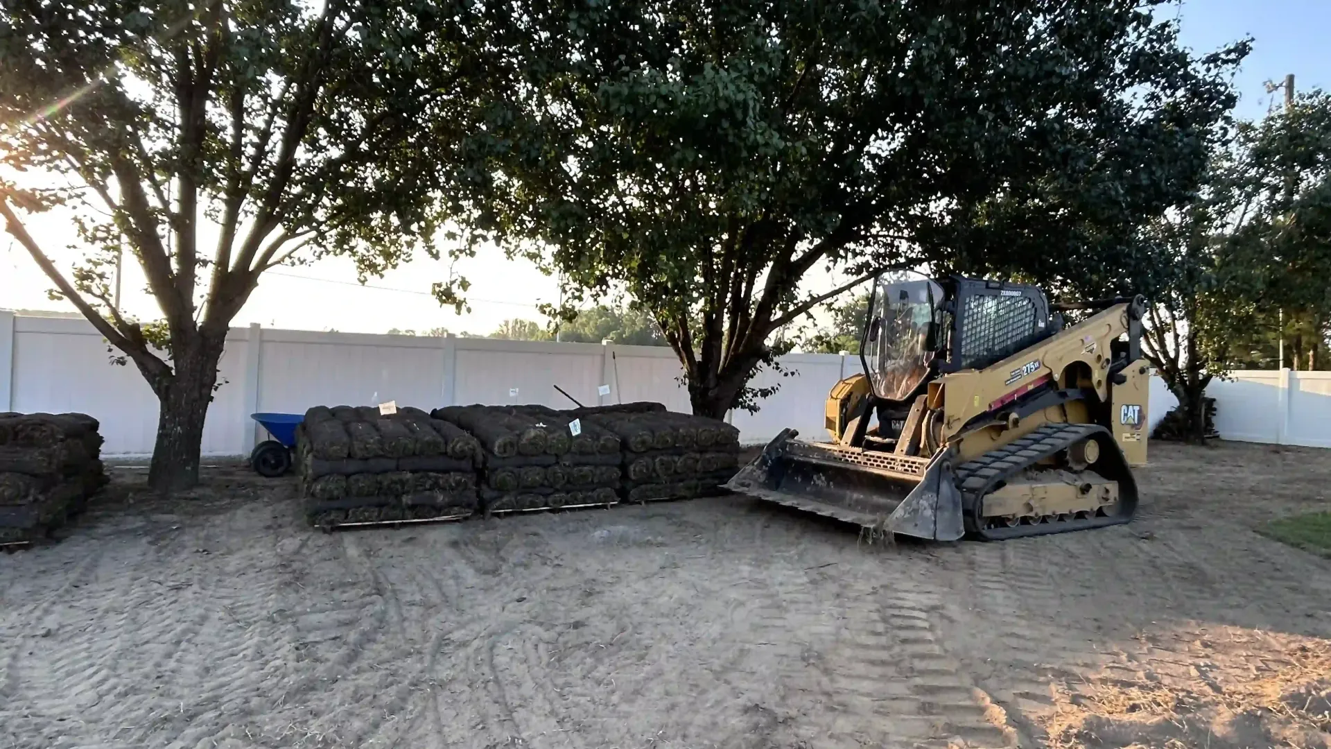Yellow skid steer clearing stacked brush piles beside a white wall under trees at dusk