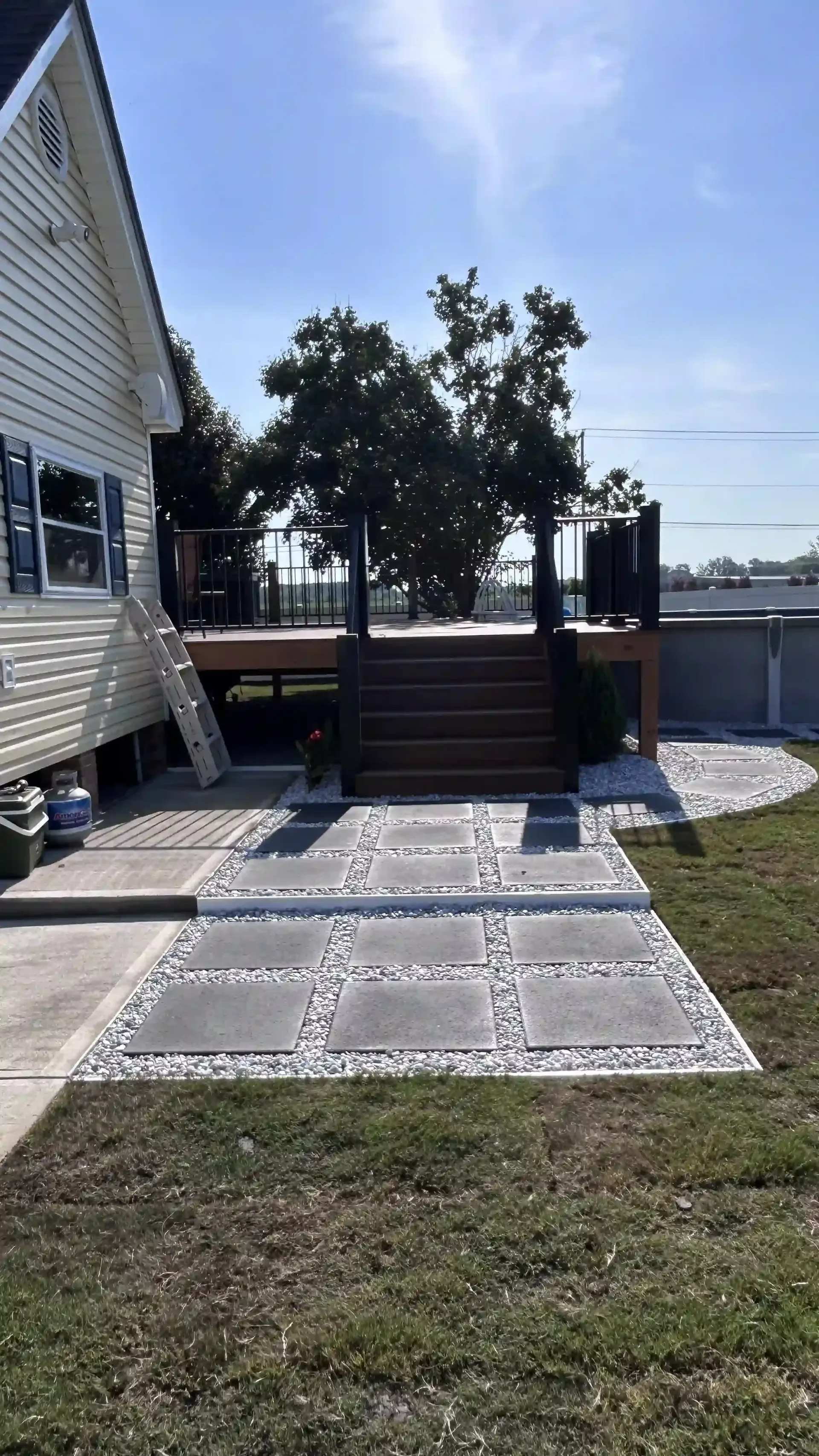 Concrete patio leading to a wooden deck beside a white house, with trees and water in the background.