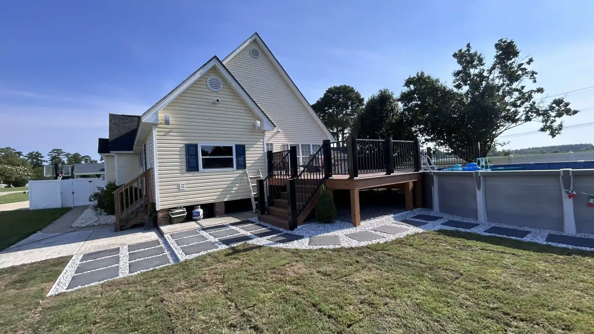 Backyard with a beige house, wooden deck, and swimming pool under a bright sky