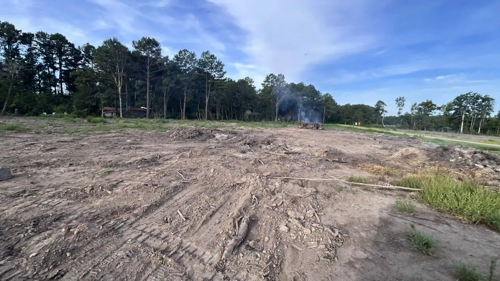 Cleared dirt lot with tire tracks, trees along the edge, and a small plume of smoke in the distance