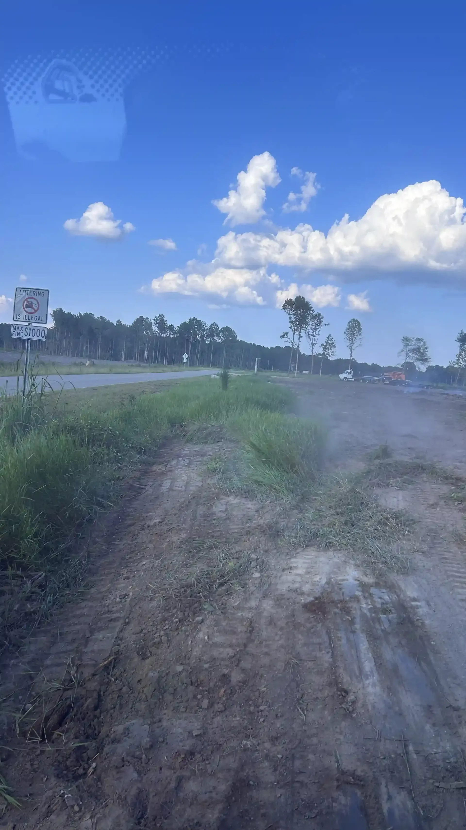 Dirt road under a bright blue sky with clouds, bordered by grass and trees.