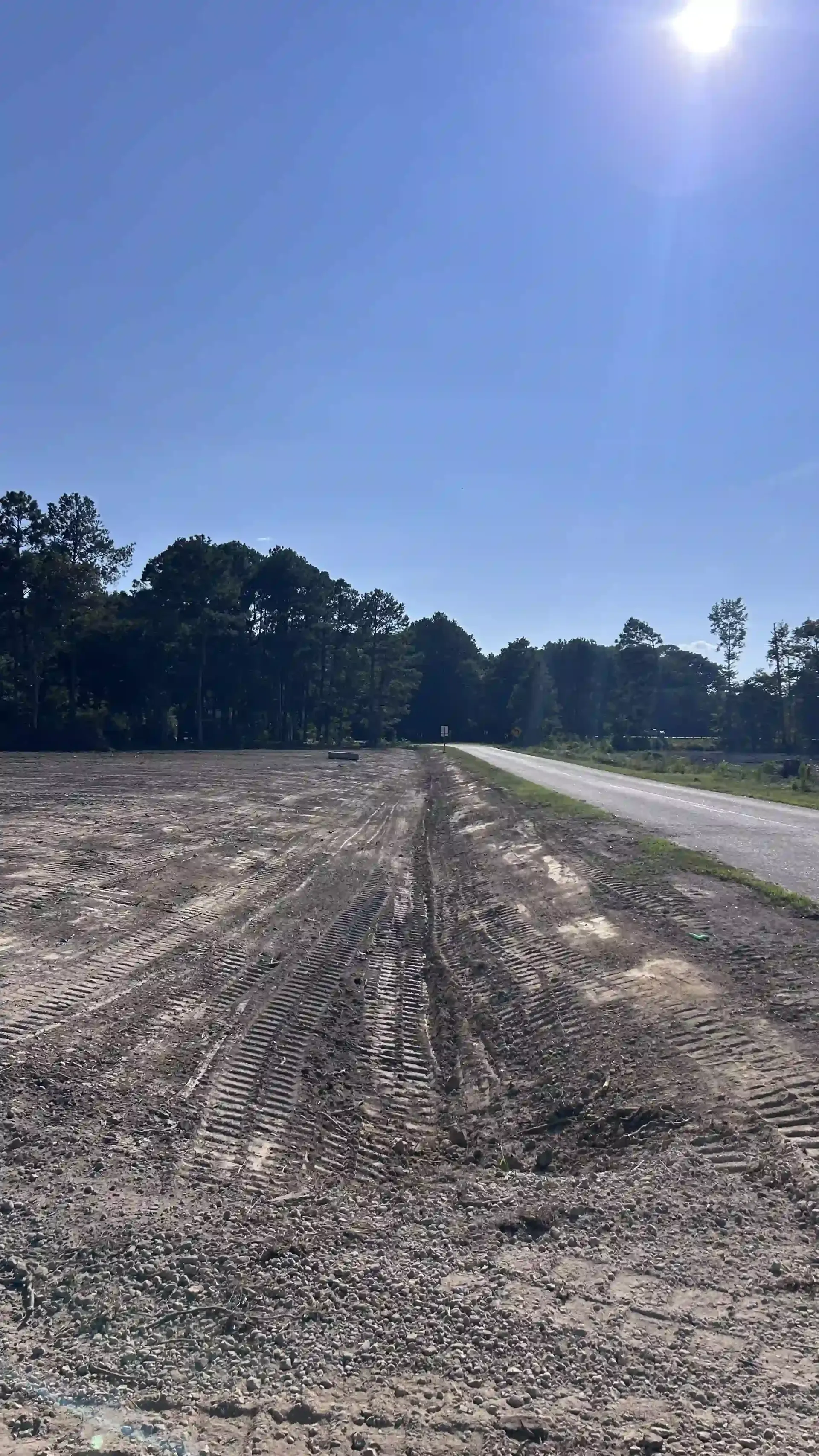 Sunlit dirt road beside a gravel lot and tree line under a clear blue sky