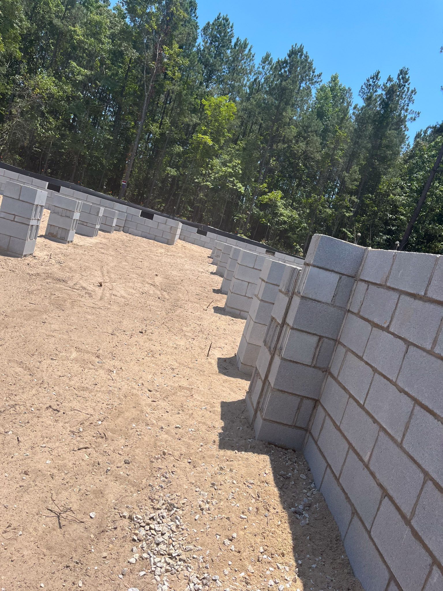 Retaining wall under construction, composed of stacked gray concrete blocks, set on a gravel base against a backdrop of green trees.