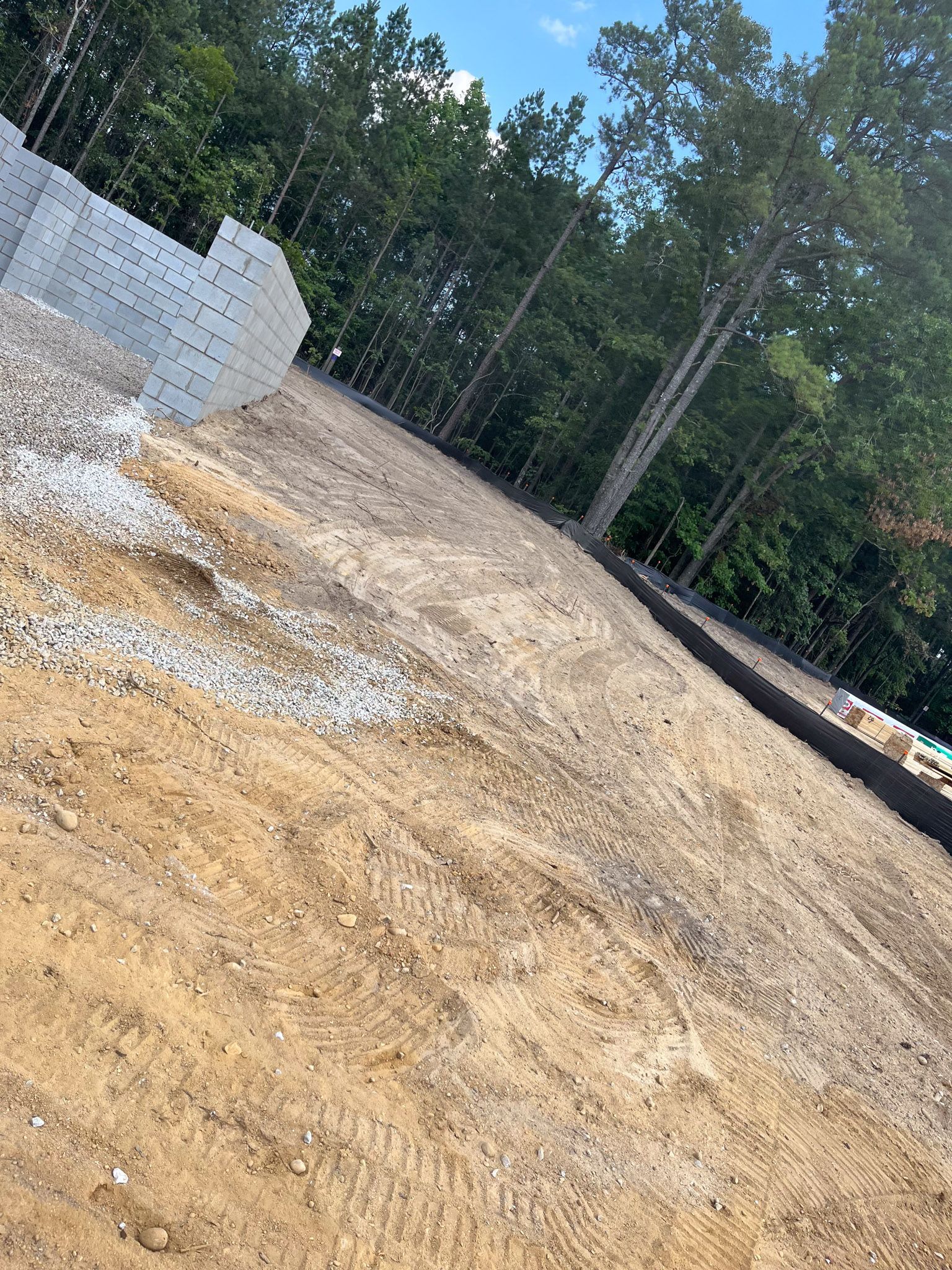Construction site with dirt, gravel, and retaining wall blocks. Trees and blue sky visible in background.