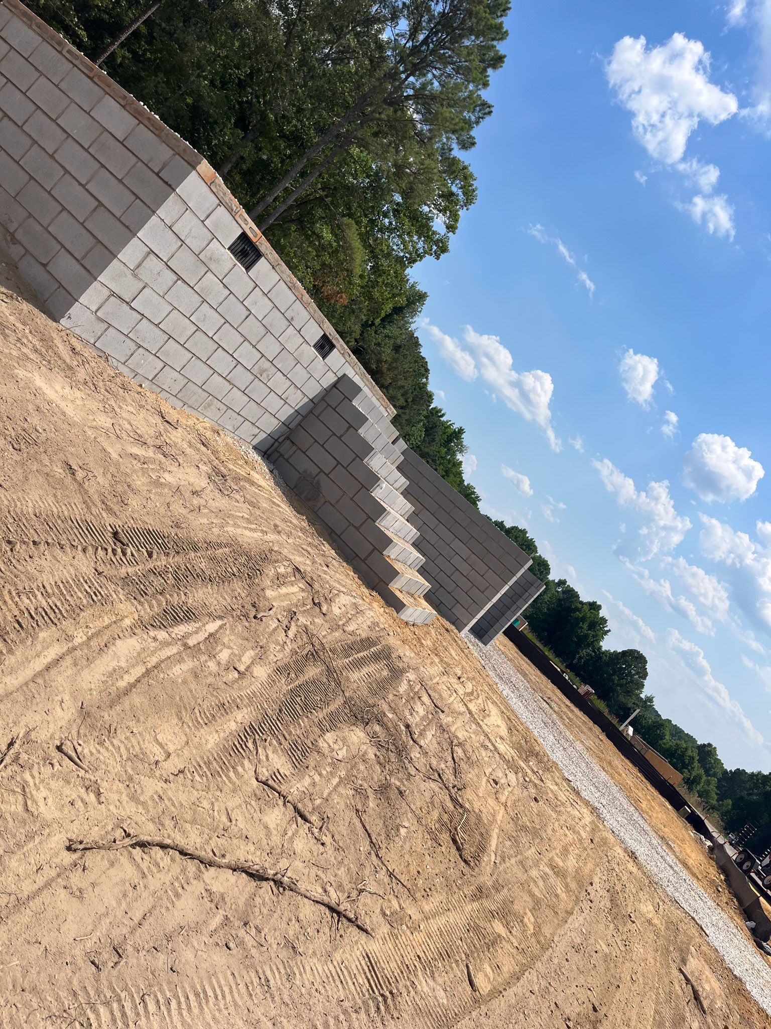 Construction site: concrete block walls being built on a graded dirt slope, under a blue sky with clouds.