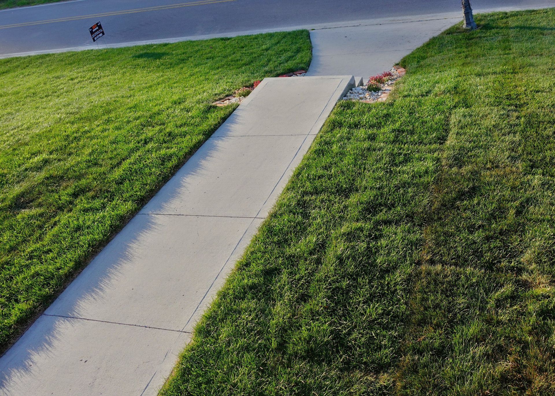 Concrete sidewalk flanked by green grass, leading to street and person on bicycle.
