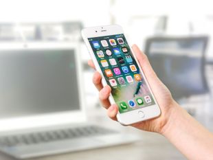 Hand holding a white smartphone displaying app icons; a laptop and office chair are in the blurred background.