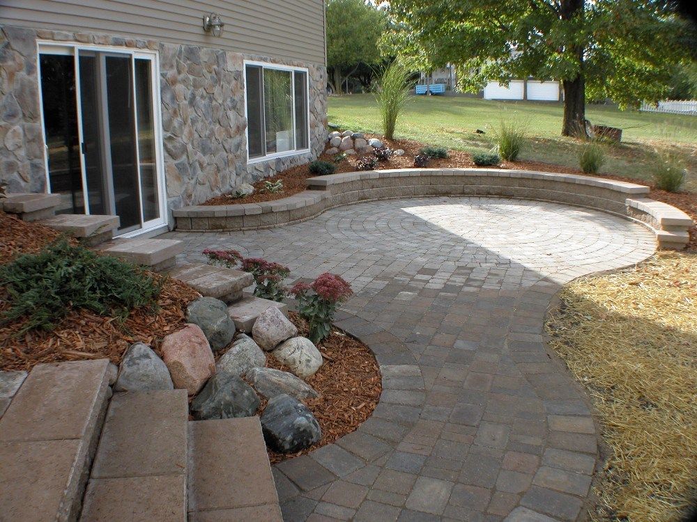 Backyard patio with brick pavers, stone retaining wall, and landscaping next to a house with sliding doors.