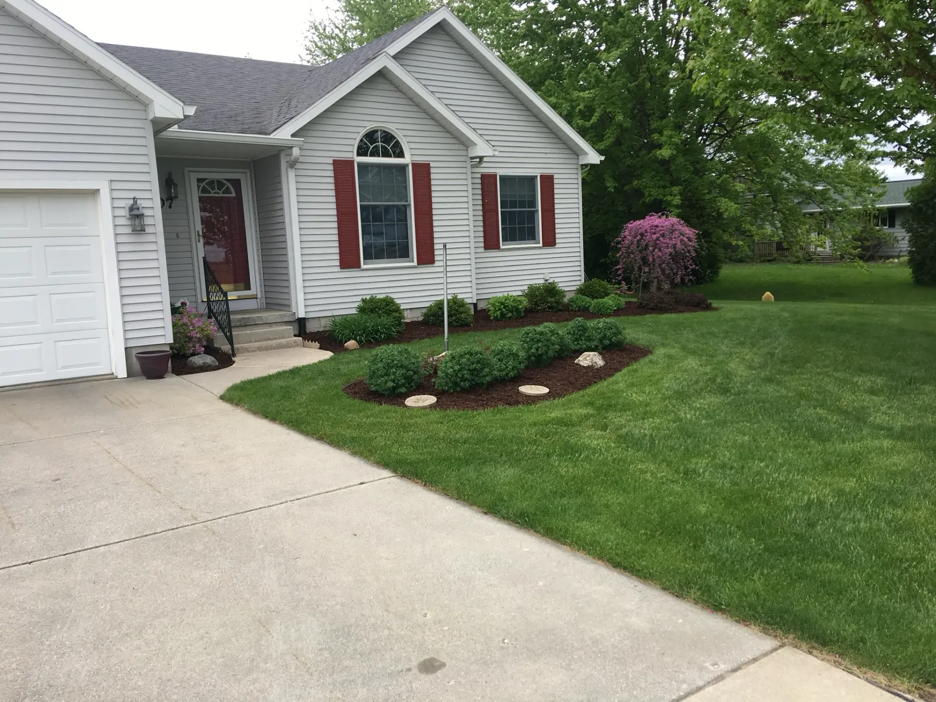 House with light gray siding, red shutters, and a flower bed on a green lawn.