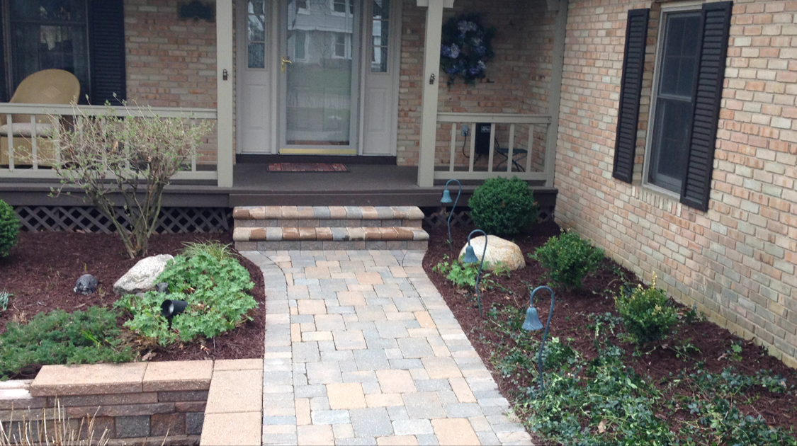 Brick home front entry with a paved walkway, landscaping, and steps leading to the door.