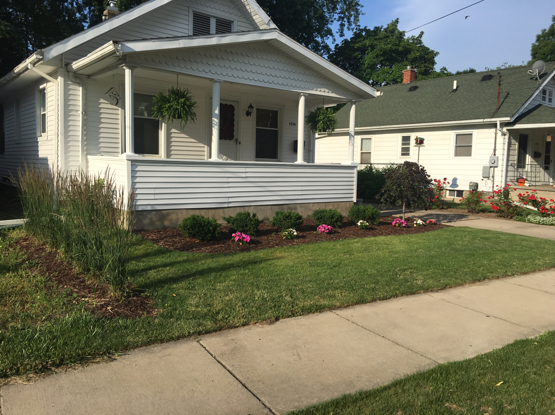 White house with a porch, flower beds, and a sidewalk on a sunny day.